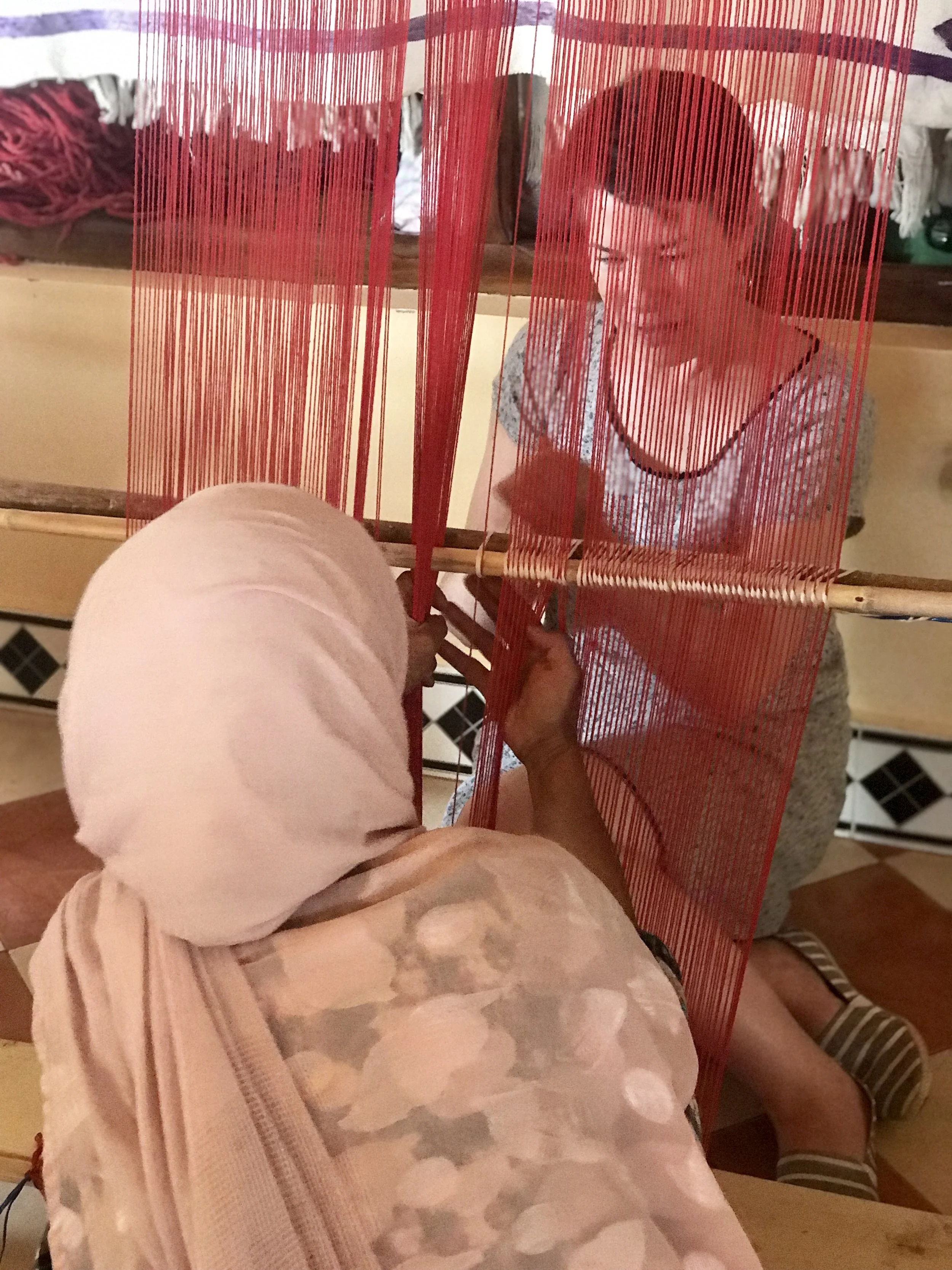 Two women are working on a red weaving loom; one is sitting on the ground, and the other is standing. They are weaving a red textile fabric. The woman sitting is wearing beige pants with a floral pattern, and the woman standing is wearing a gray dress with black and white striped shoes.