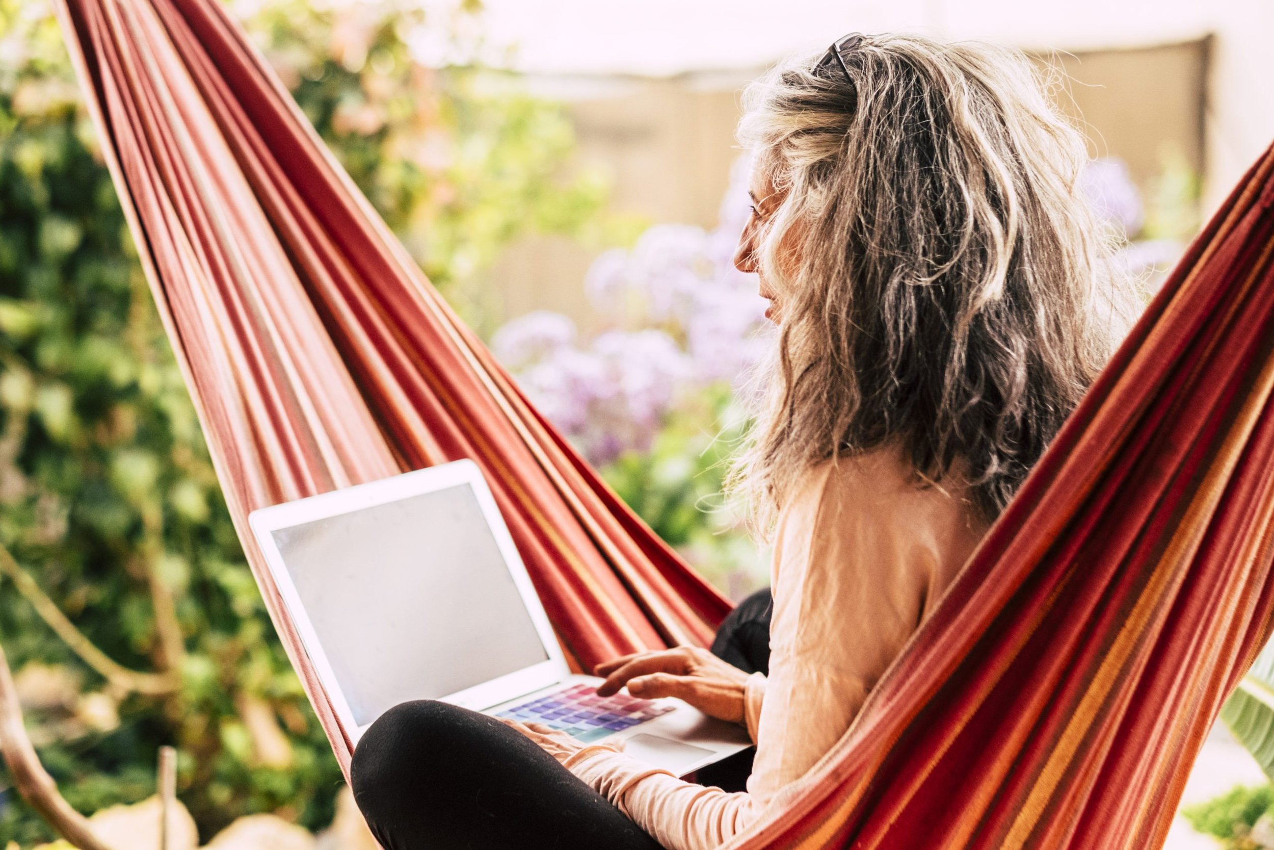 A woman with gray and black curly hair sitting in a hammock outdoors, using a laptop, with a background of greenery and flowers.