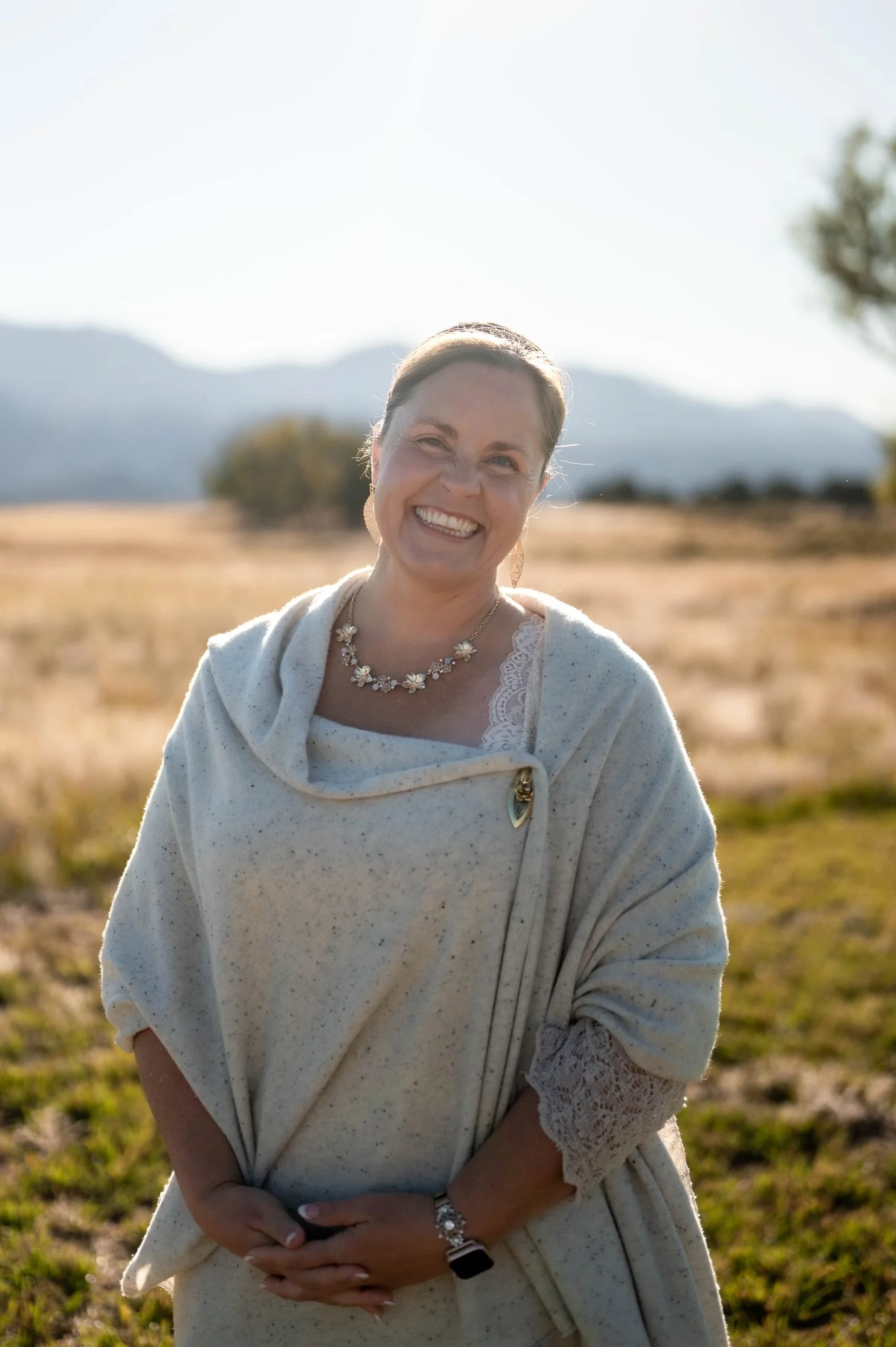 A smiling woman standing outdoors in a field with mountains in the background, wearing a light-colored sweater and jewelry, backlit by the sun.