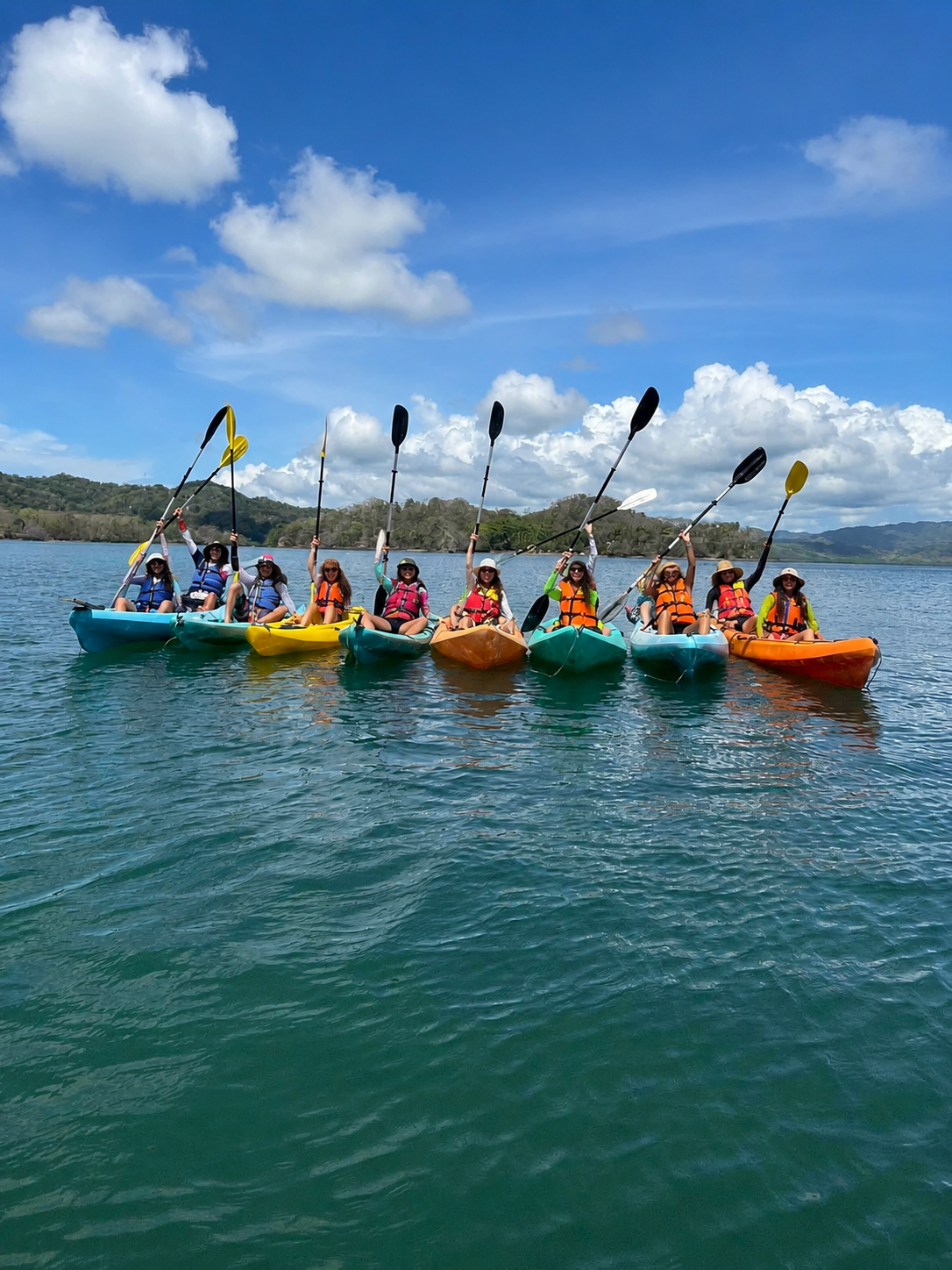 Multiple yellow kayaks with people paddling near a lush green island surrounded by blue water under a partly cloudy sky.