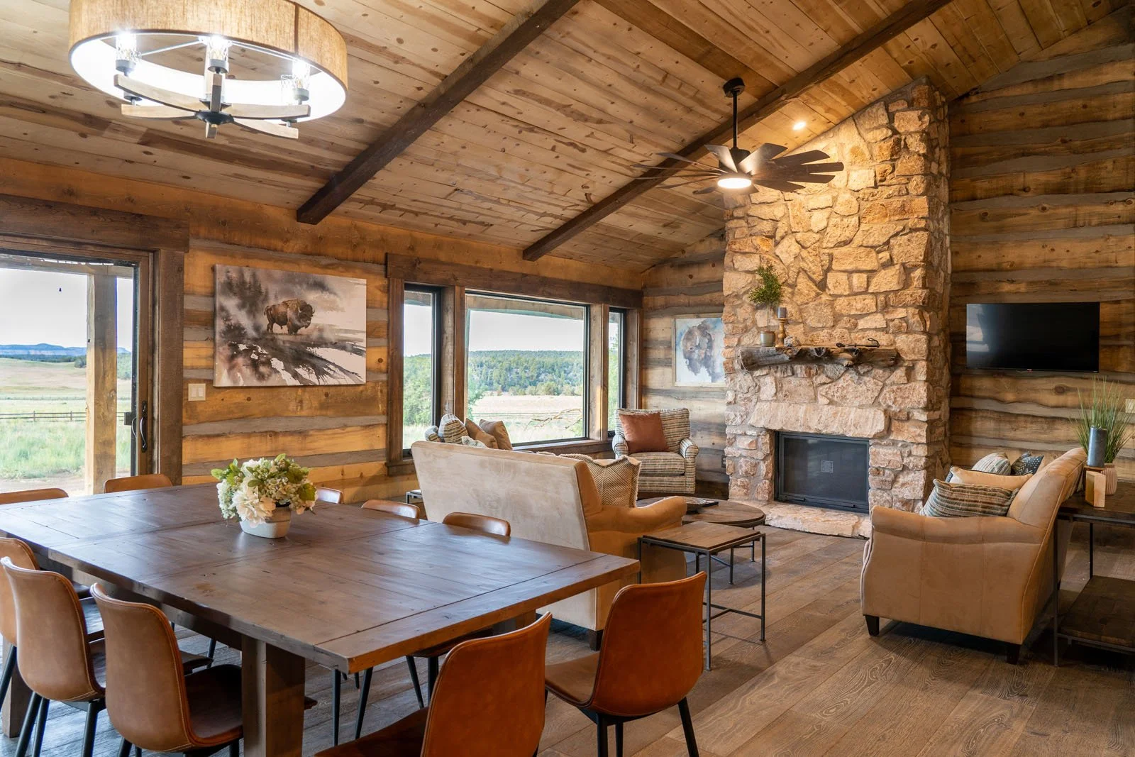 Interior of a rustic living room with wood-paneled walls and ceiling, a stone fireplace, a mounted TV, large windows showing a scenic landscape, and various seating including a dining table, armchairs, and a sofa.