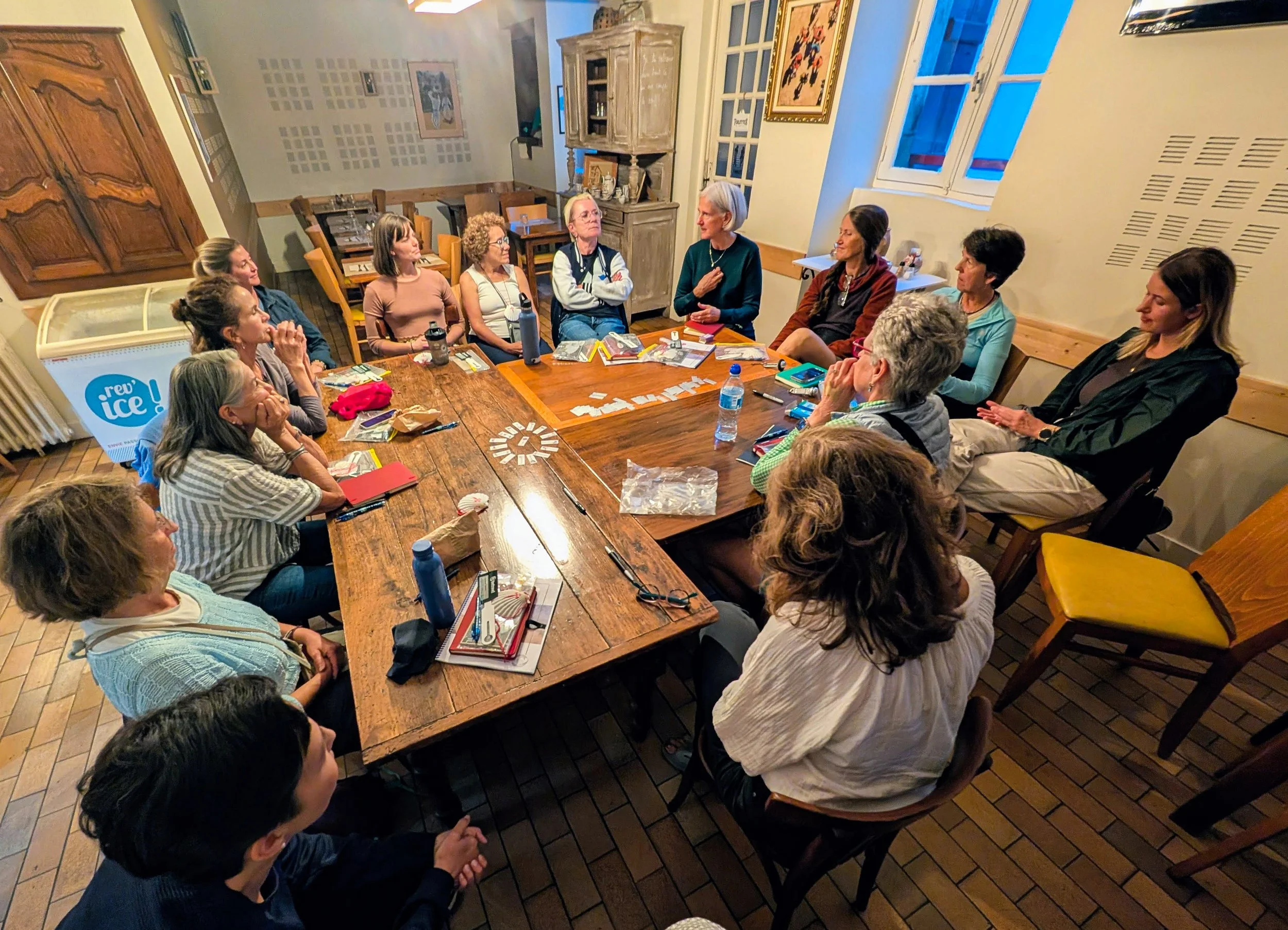 Group of women sitting around a large wooden table in a cozy room, engaged in a discussion. Some have notebooks, water bottles, and other items in front of them.