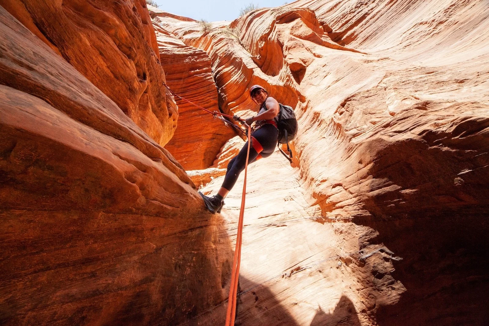 A person wearing a helmet, sunglasses, and outdoor gear is rappelling down a narrow canyon with reddish-orange sandstone walls.