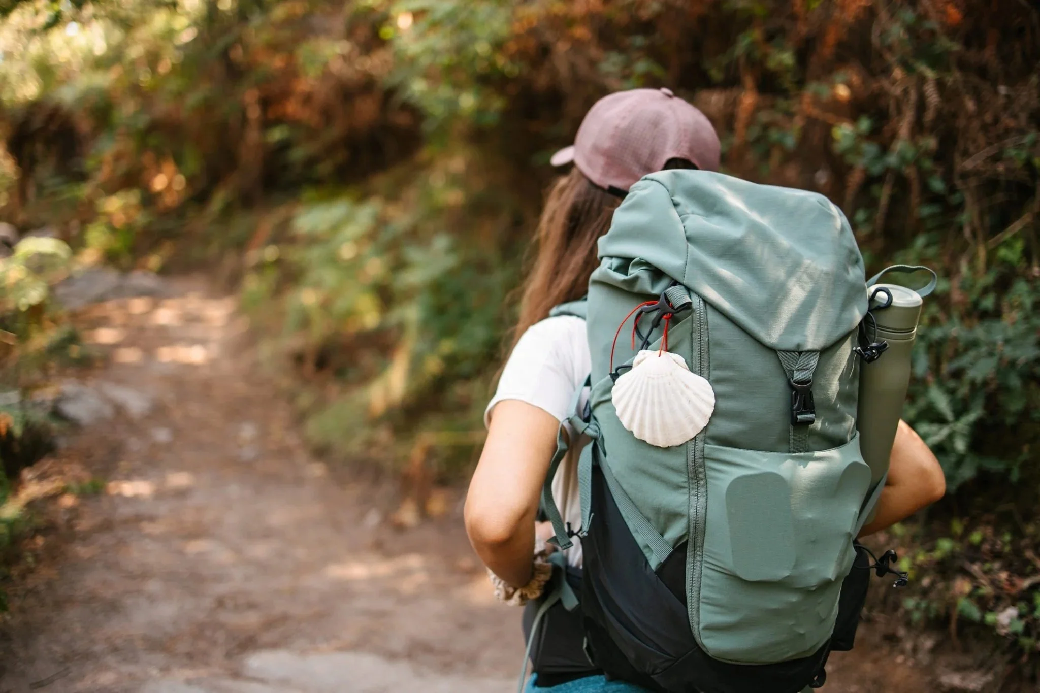 A person hiking on a trail through a forest, wearing a pink cap, and carrying a large backpack with a seashell attached.