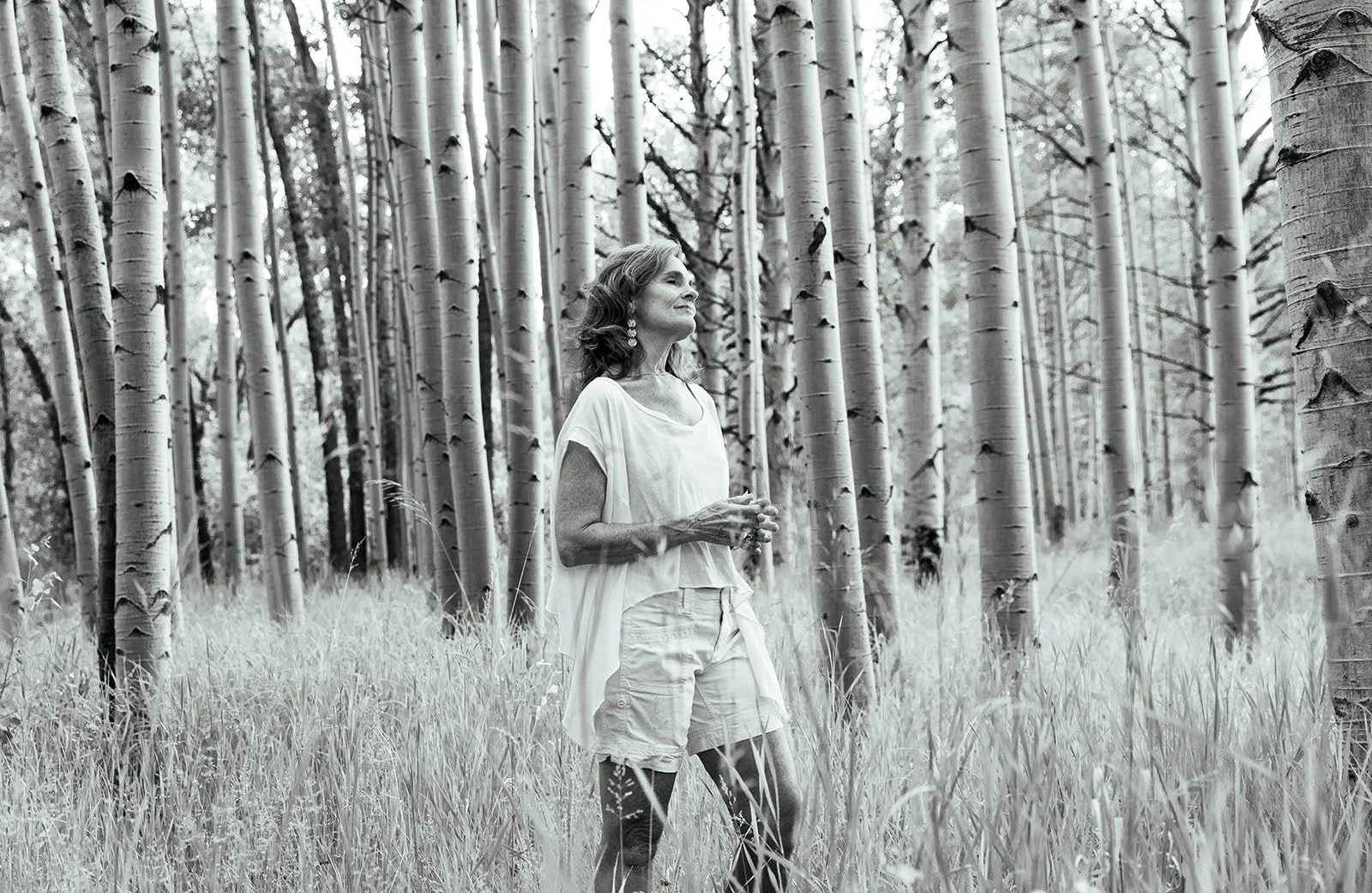 A woman stands amidst tall grass and trees in a forest, looking thoughtfully into the distance.