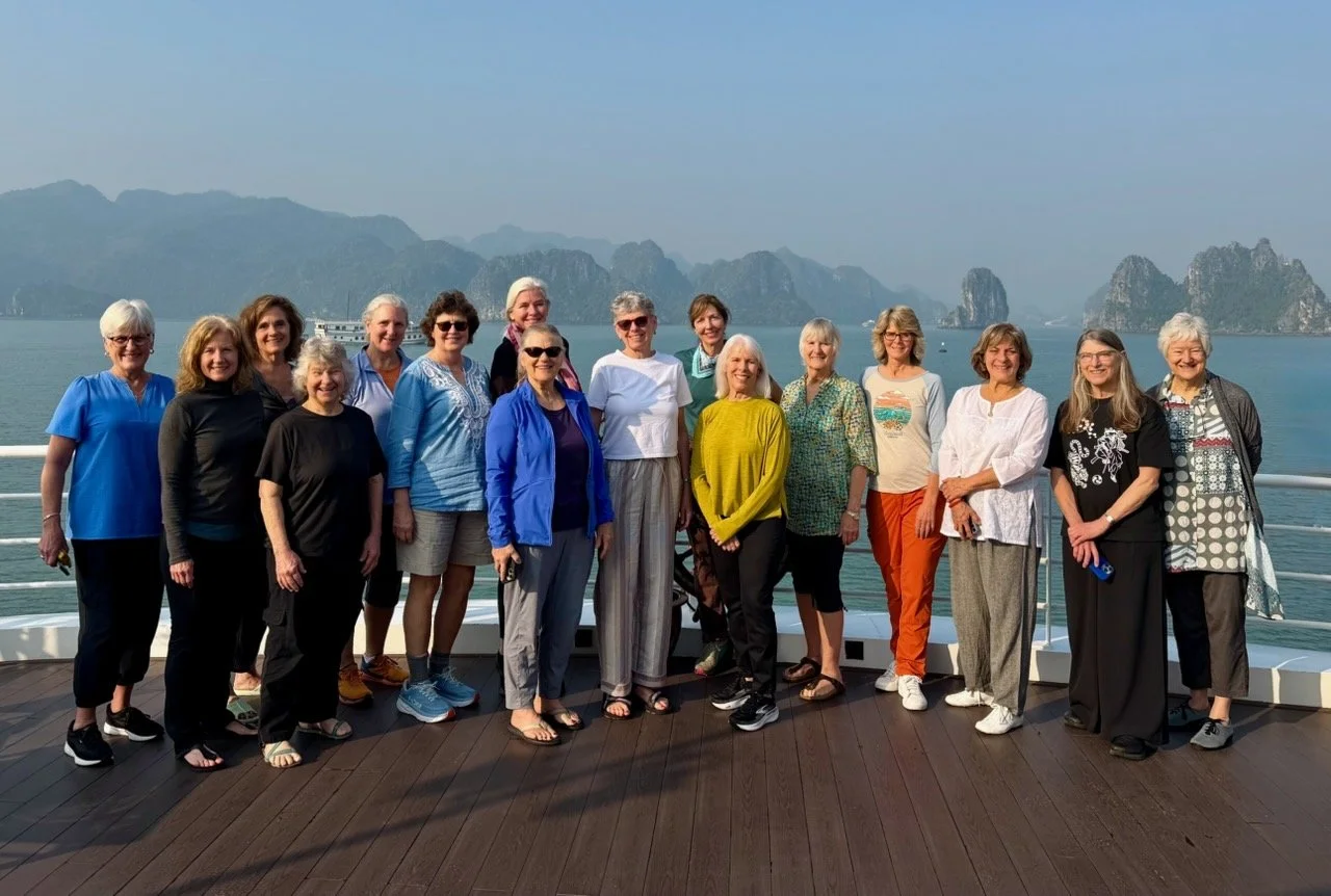 A group of 15 women standing on the deck of a boat with a scenic bay landscape in the background, including karst limestone formations and calm water.