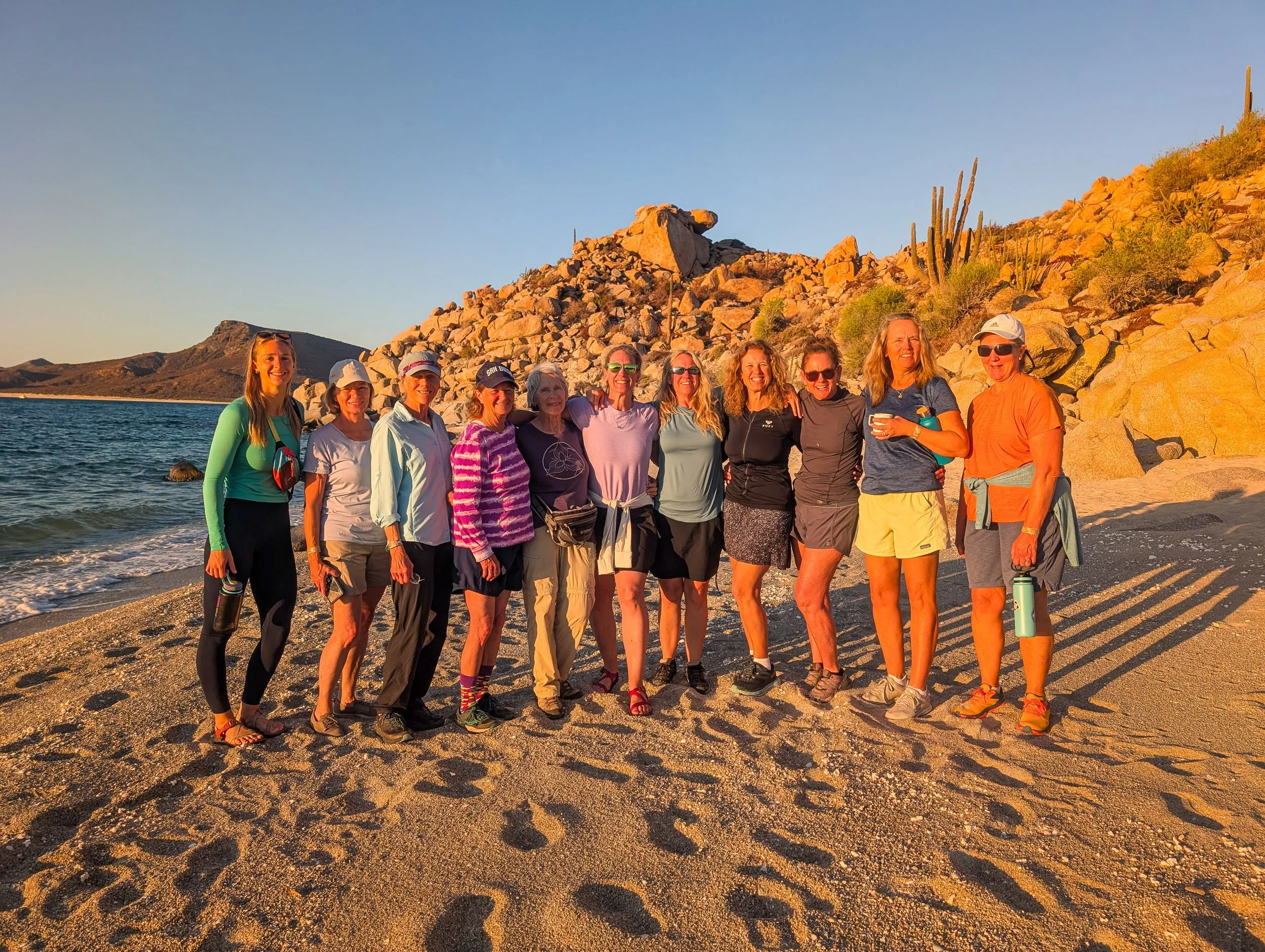 Group of twelve women standing on a beach with rocks and cacti in the background, during sunset.