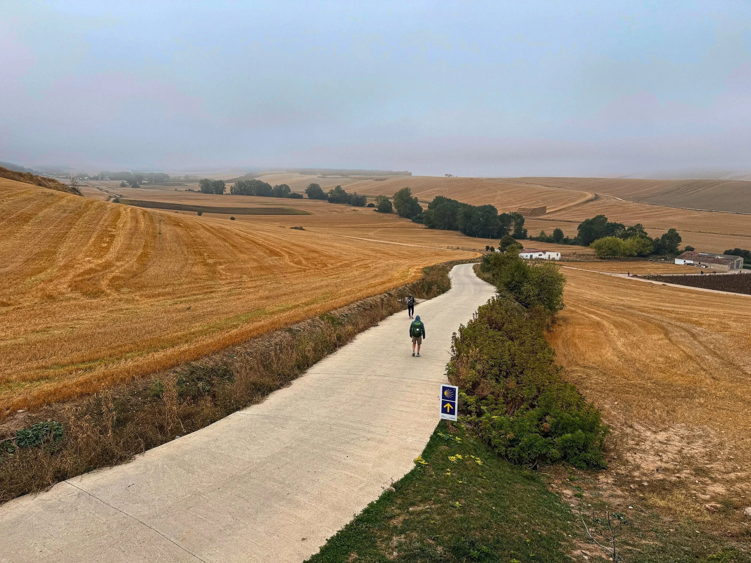 Two hikers walking along the El Camino de Santiago trail in a rural landscape with golden fields, distant farm buildings, and a cloudy sky.