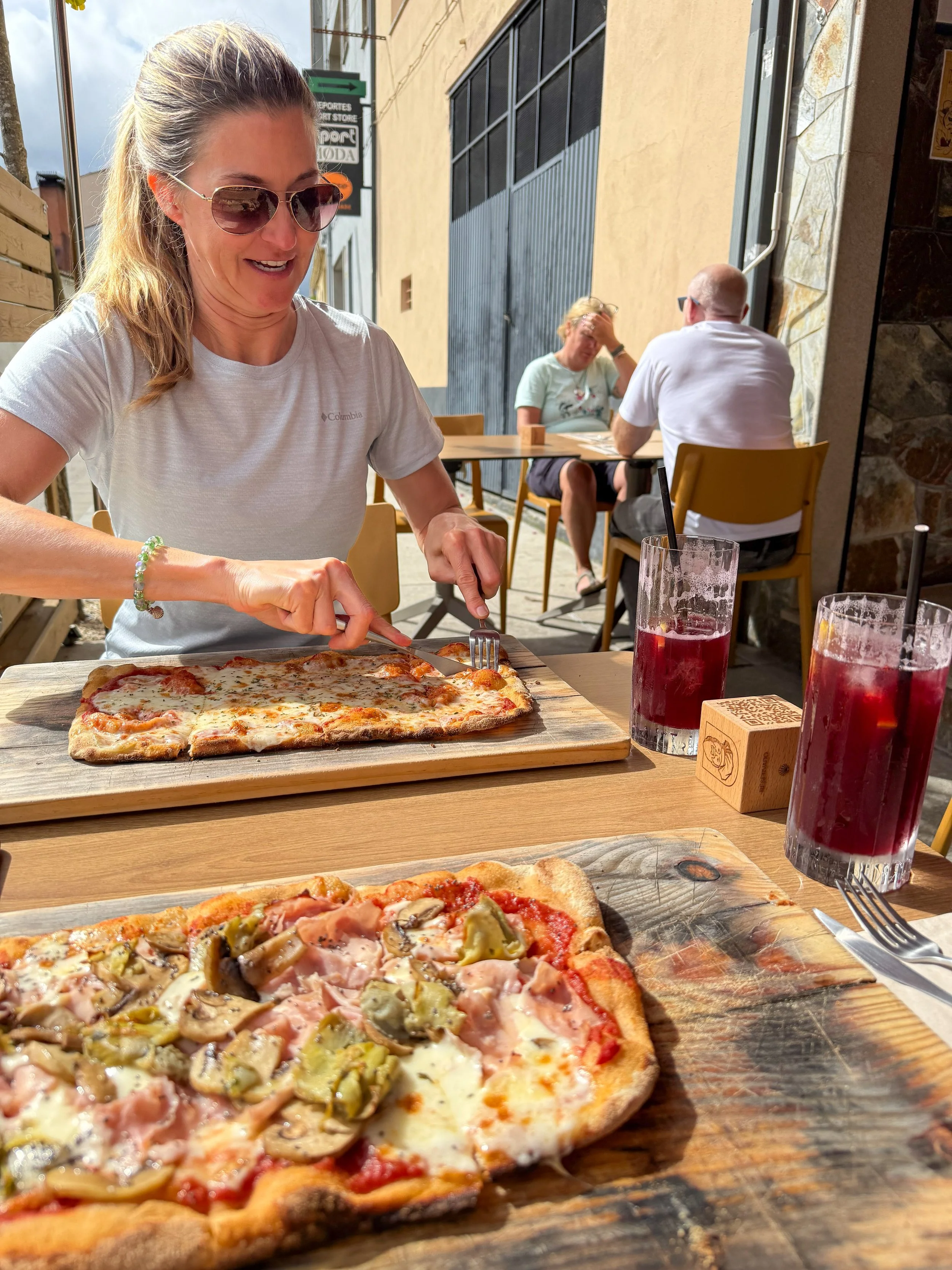 A woman sitting at an outdoor restaurant table cutting into a rectangular cheese pizza with a woman and a man sitting behind her at a table. There are two glasses of dark red beverages on the table.