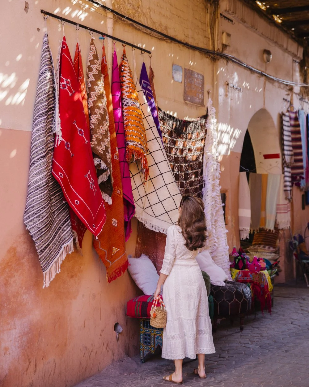 A woman in a white dress shopping for colorful patterned rugs displayed on a wall in a marketplace.