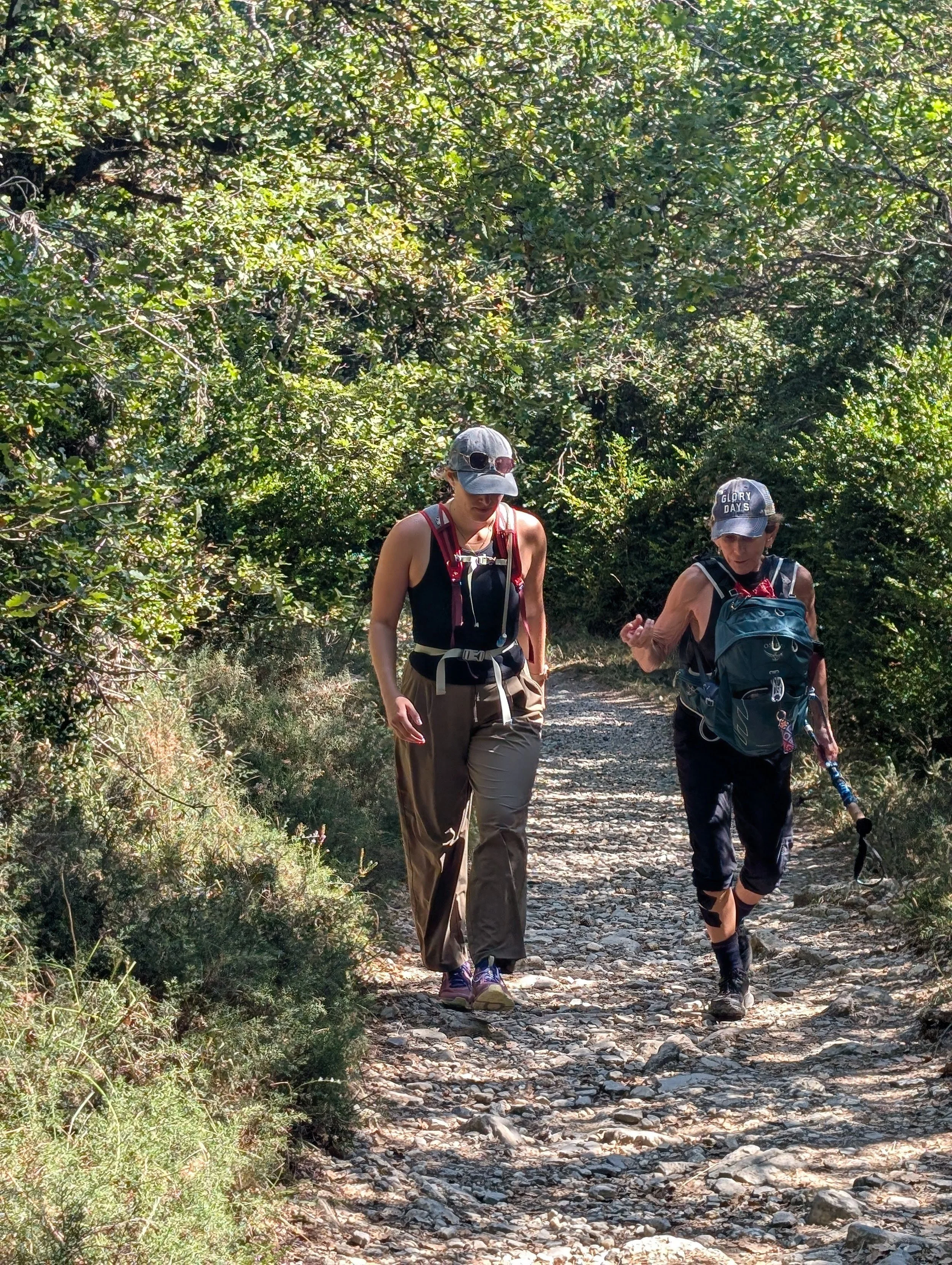 Two women trail walking on a rocky dirt path surrounded by dense green foliage and bushes, wearing hats, backpacks, and outdoor gear.