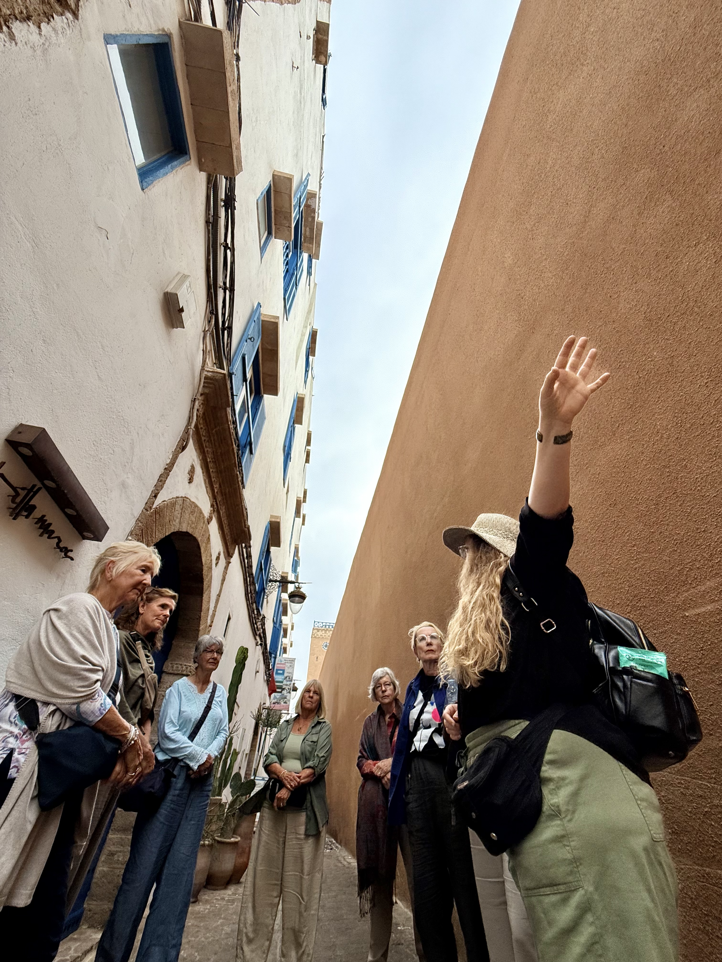 Tour group standing on a narrow street surrounded by colorful buildings, some with blue window frames, listening to a guide, while one woman with long blonde hair and a hat gestures with her hand.