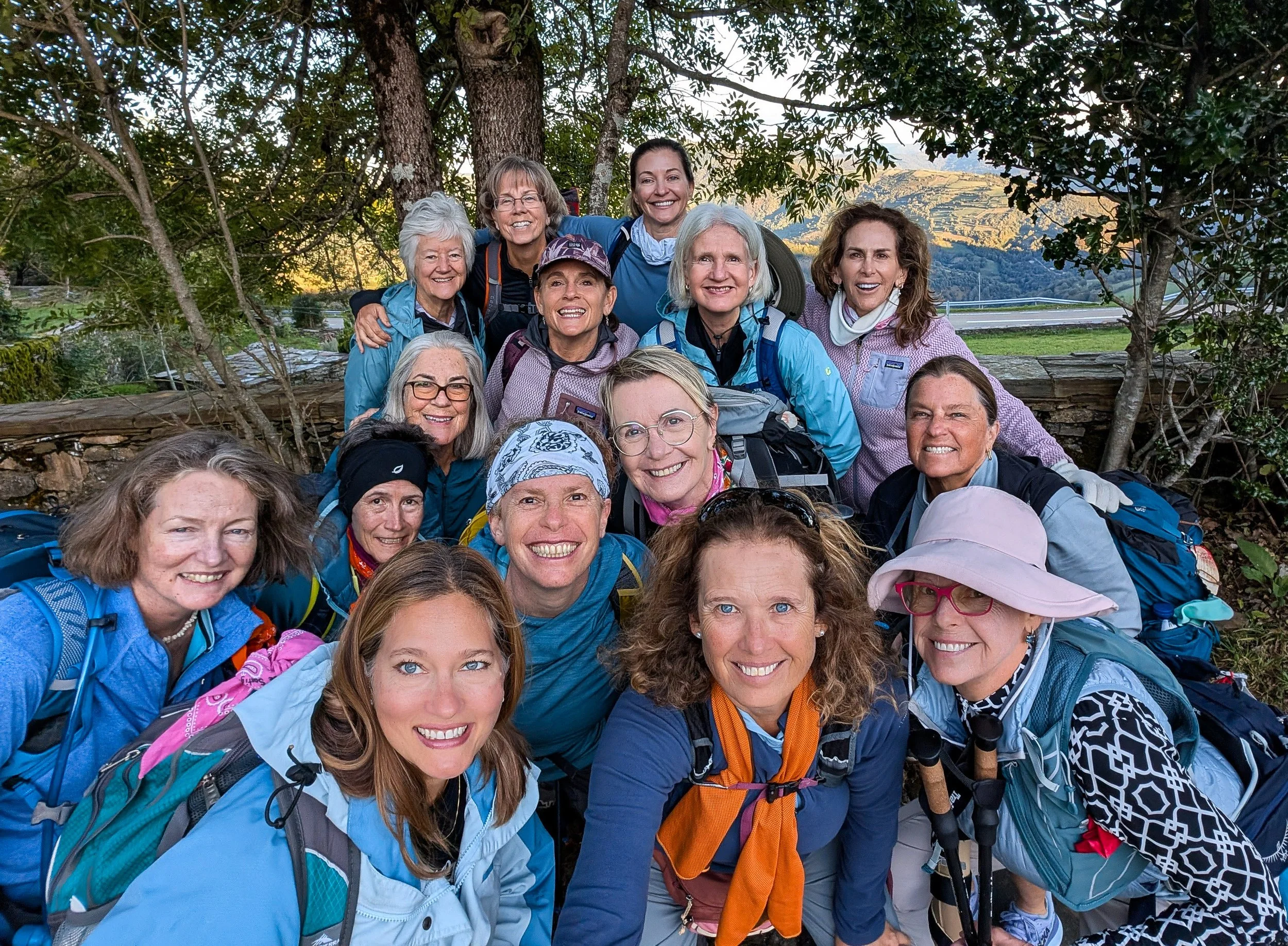 Group of 15 women smiling for a photo outdoors during a hike, wearing outdoor gear and backpacks, with trees and mountains in the background.