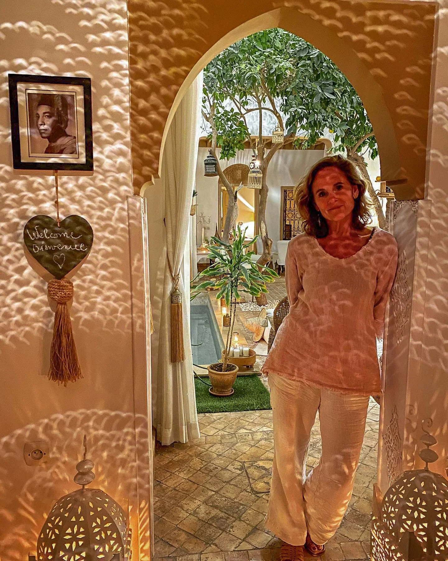 A woman standing in a warmly lit, cozy indoor space with Moroccan-inspired decor, including decorative lanterns and a mirror creating light patterns on the wall. She is smiling and dressed in light-colored pajamas, with a green potted plant and a tree with hanging lanterns visible in the background.