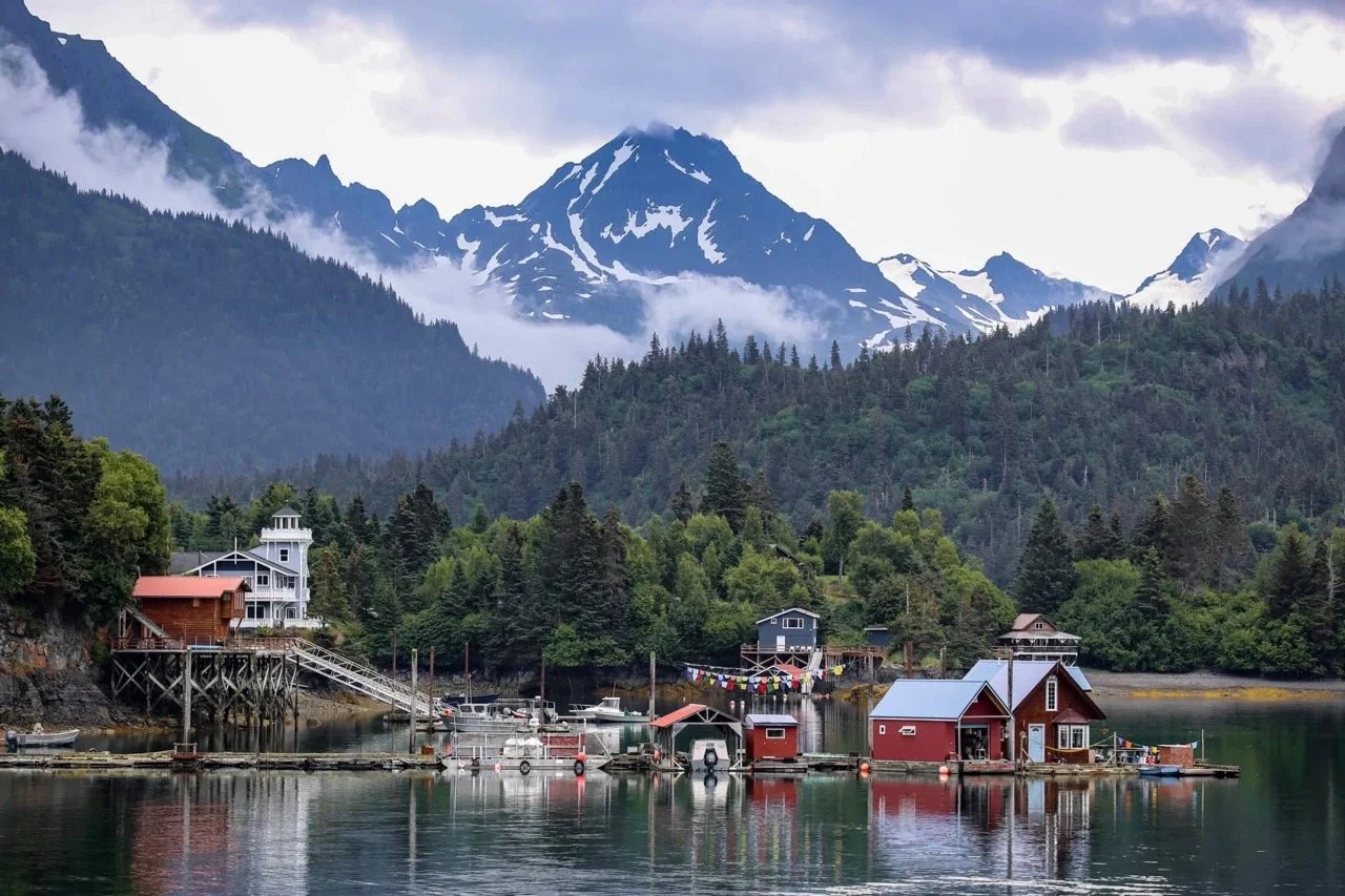 Alaska in august scenic view of a turquoise lake surrounded by pine trees and tall mountains with patches of snow under a partly cloudy sky.