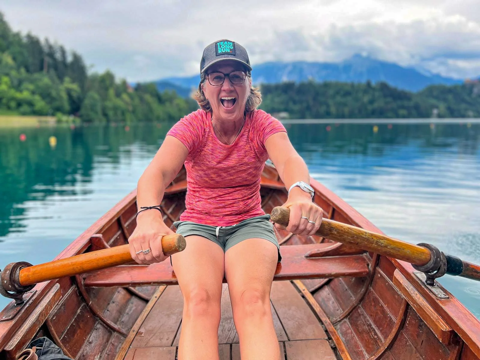 A woman in a pink shirt and shorts rowing a boat on a lake with green trees and mountains in the background.