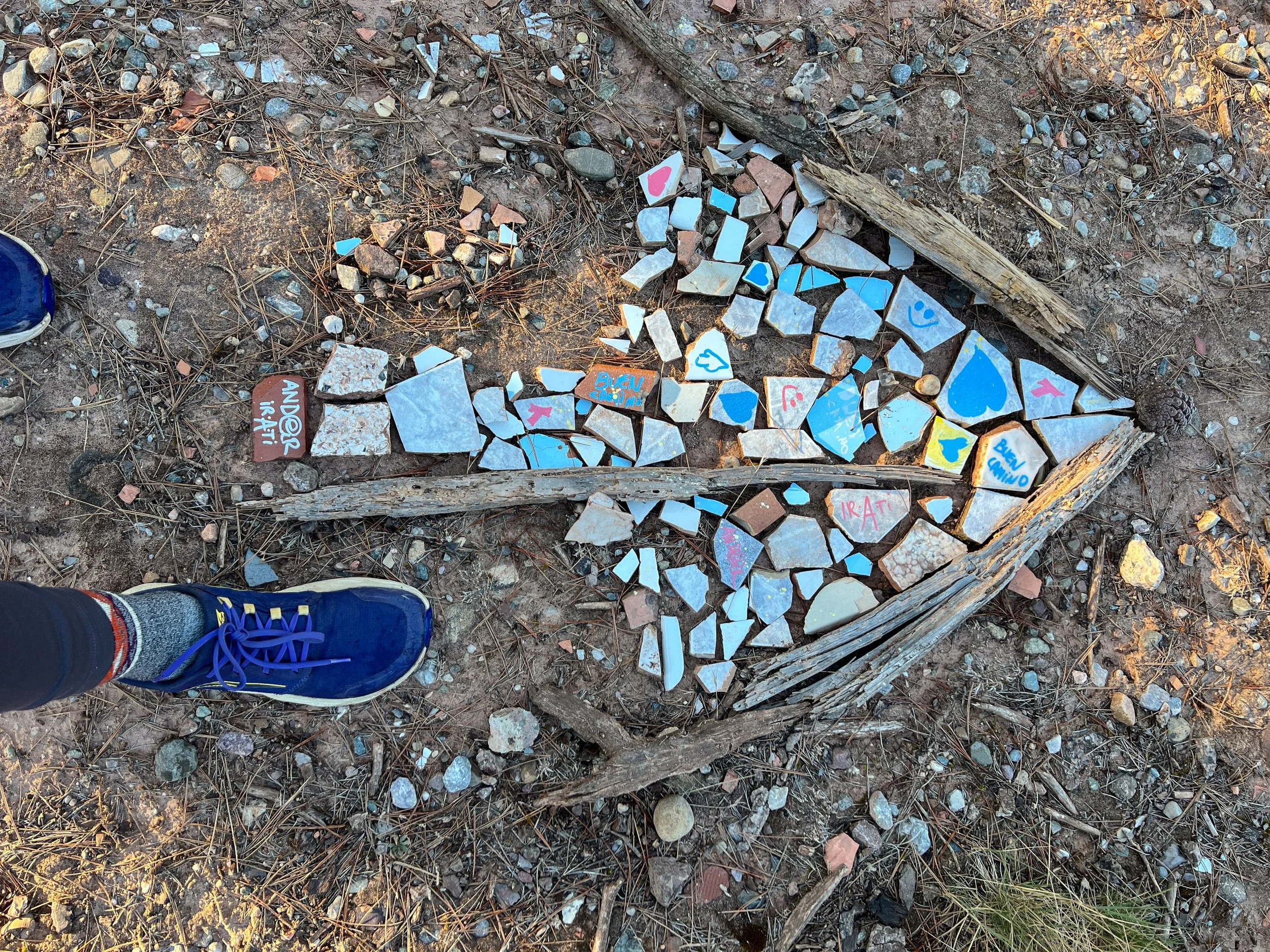 Broken tiles arranged in a triangular shape on the ground, surrounded by dirt, sticks, and small rocks; one tile has artistic writing and colorful designs; a person's feet and shoes are visible at the bottom.