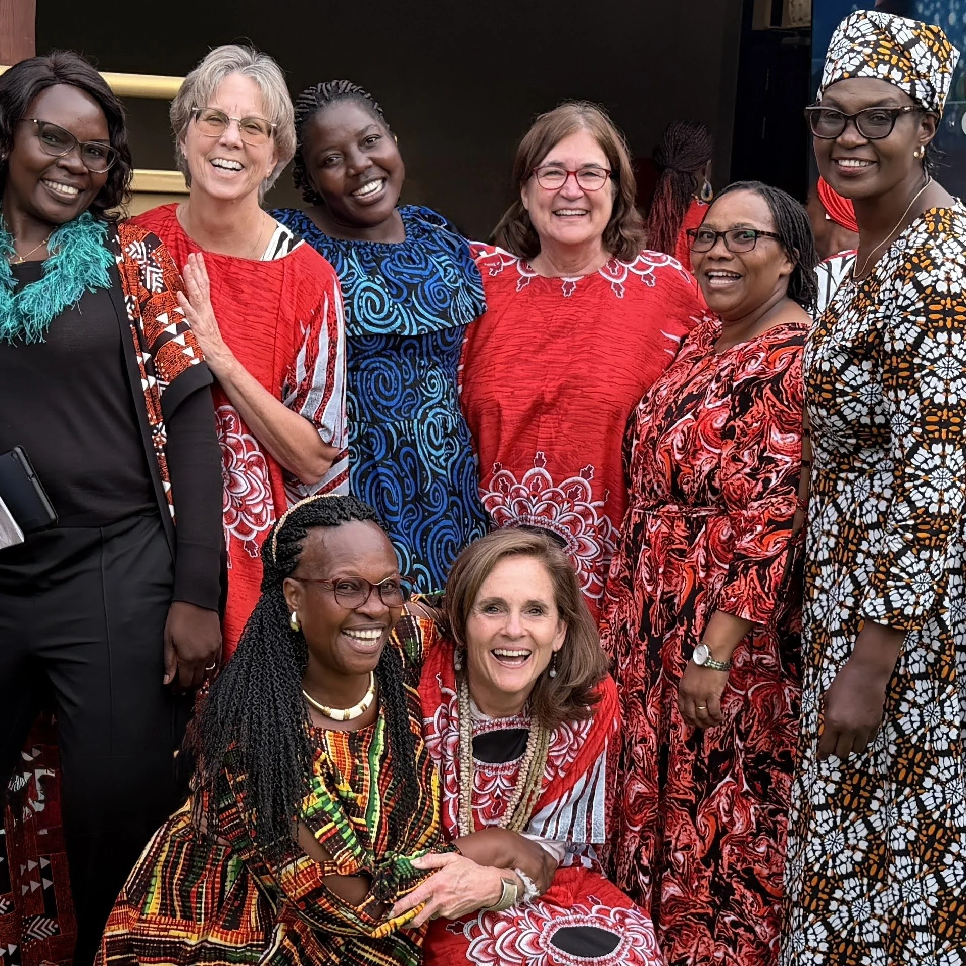 Group of women from diverse backgrounds wearing traditional and colorful clothing, smiling and posing together at a social gathering.