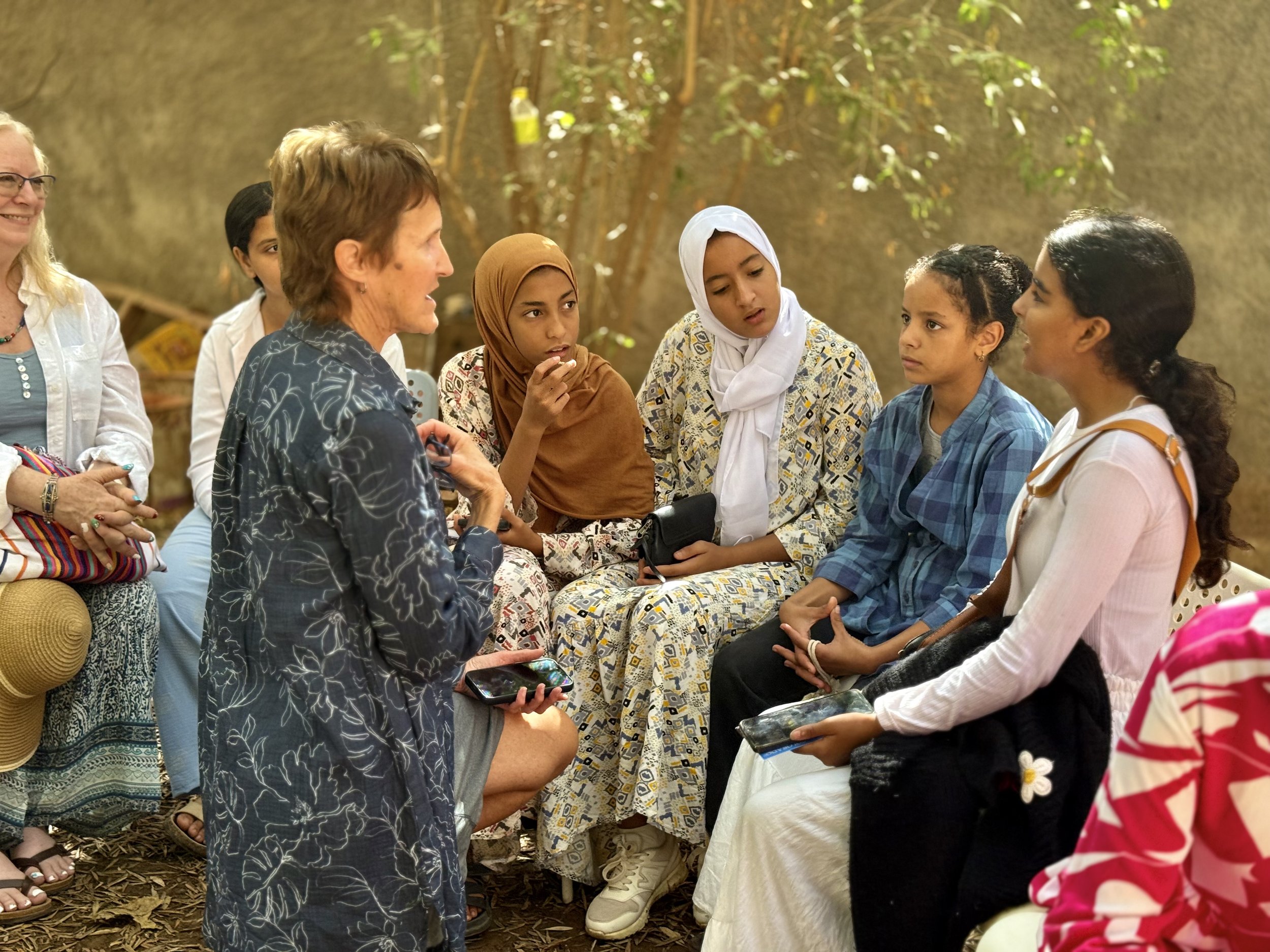 A group of ethnically diverse women and girls engaged in a discussion outdoors, with trees and natural surroundings in the background.