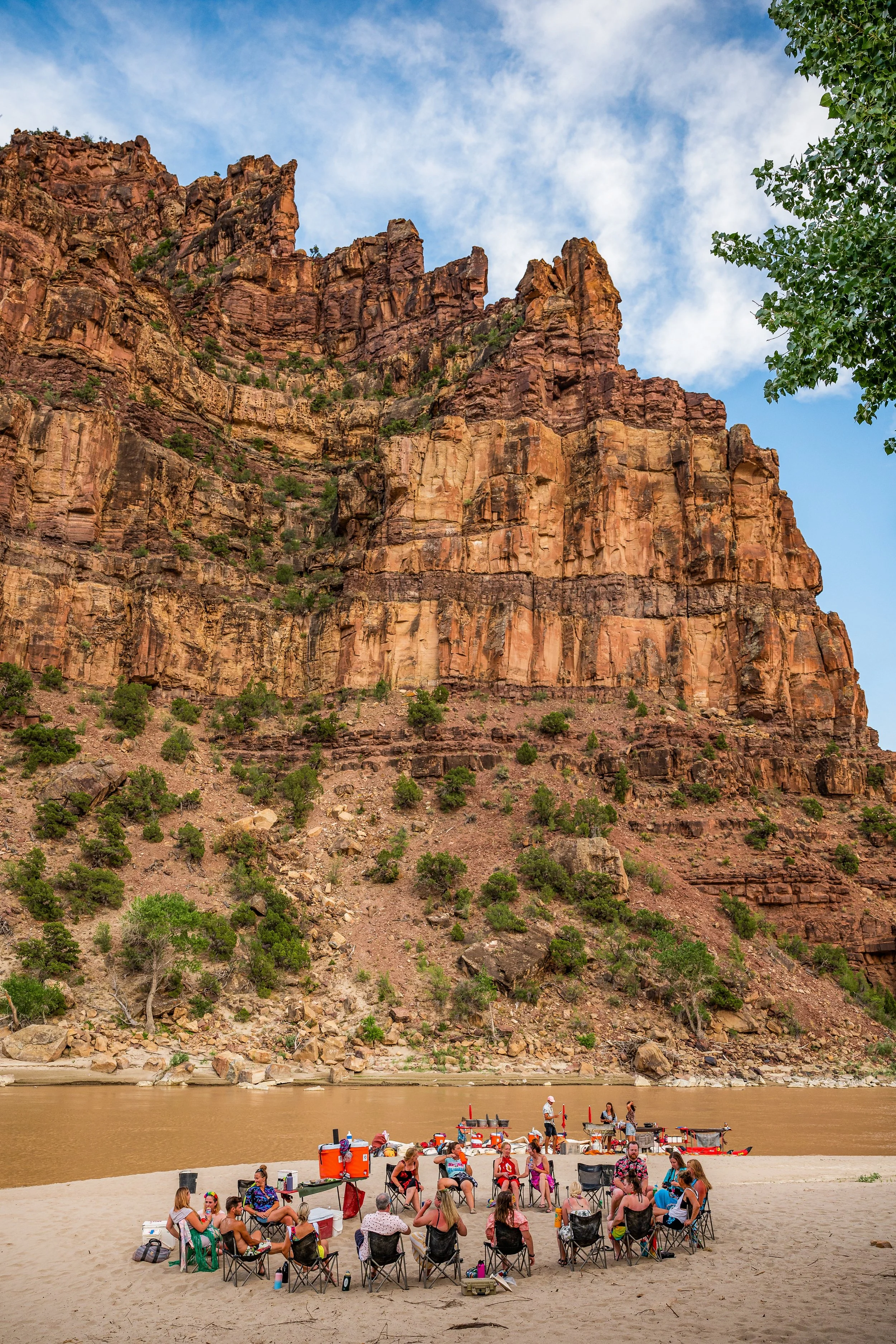 A group of smiling people wearing sunglasses and life jackets, paddling in a line in a river with red kayaks in the background.