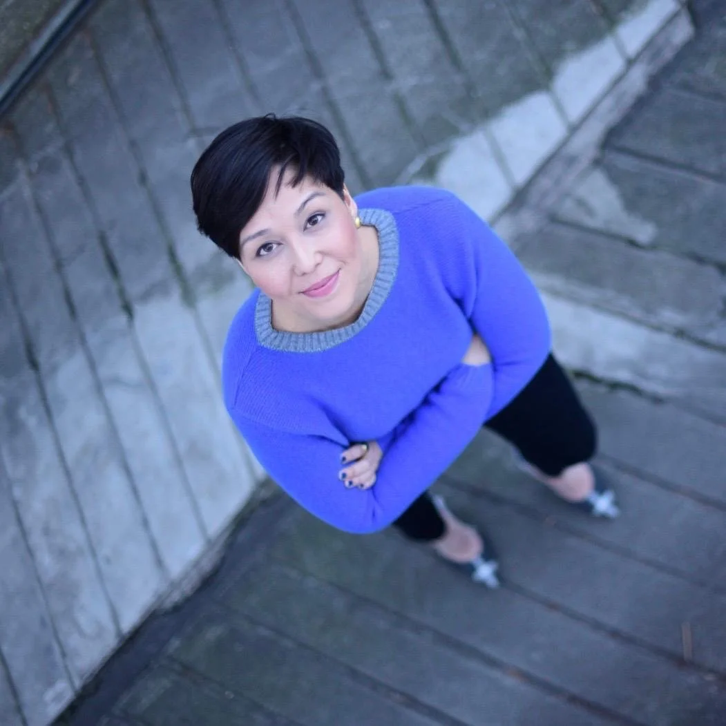 A woman with short dark hair standing on a wooden deck and posing for the camera, wearing a blue sweater with gray collar/details, black pants, and black and white shoes, with a stone wall in the background.