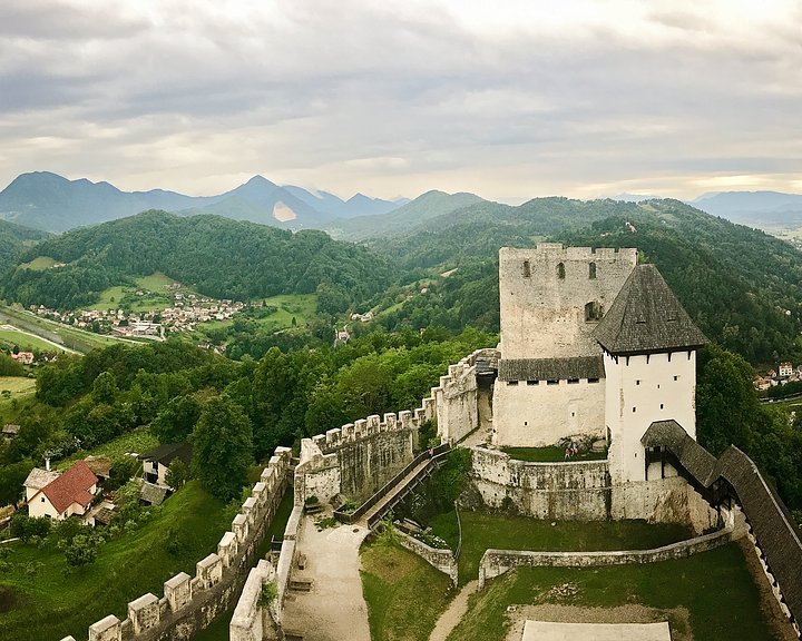 A medieval castle with white walls and black accents, surrounded by green trees and hills, under a cloudy sky.