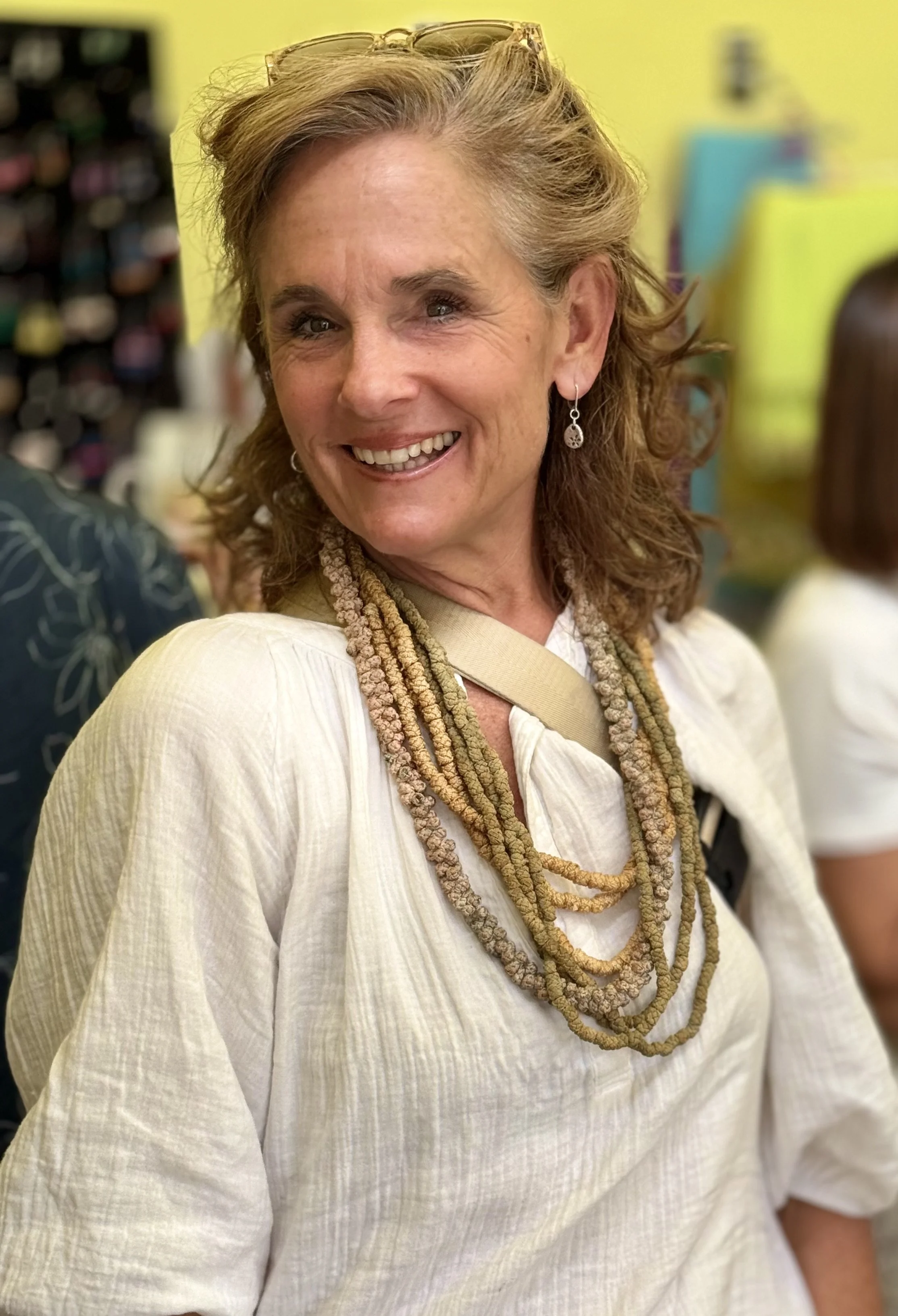 Close-up of a smiling woman's face with light brown, wavy hair, wearing glasses on her head, earrings, and layered necklaces, in an indoor setting with yellow and colorful background.