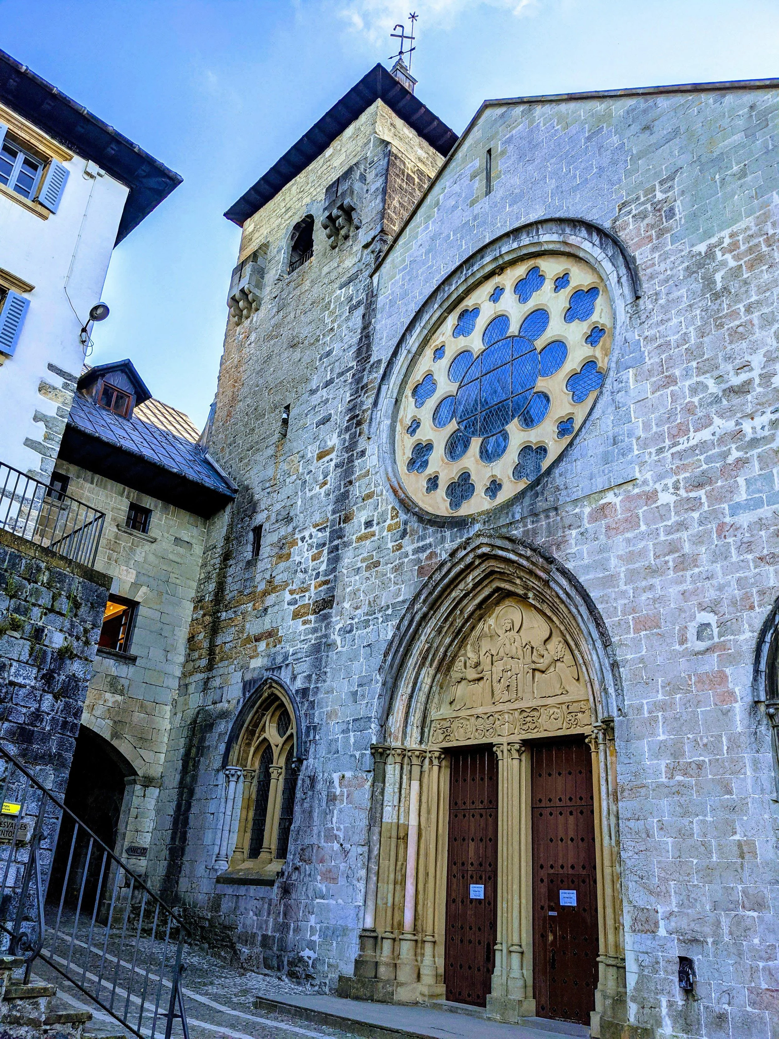 The exterior of an old stone church with a large circular stained glass window and a carved wooden entrance door, located on a narrow cobblestone street.