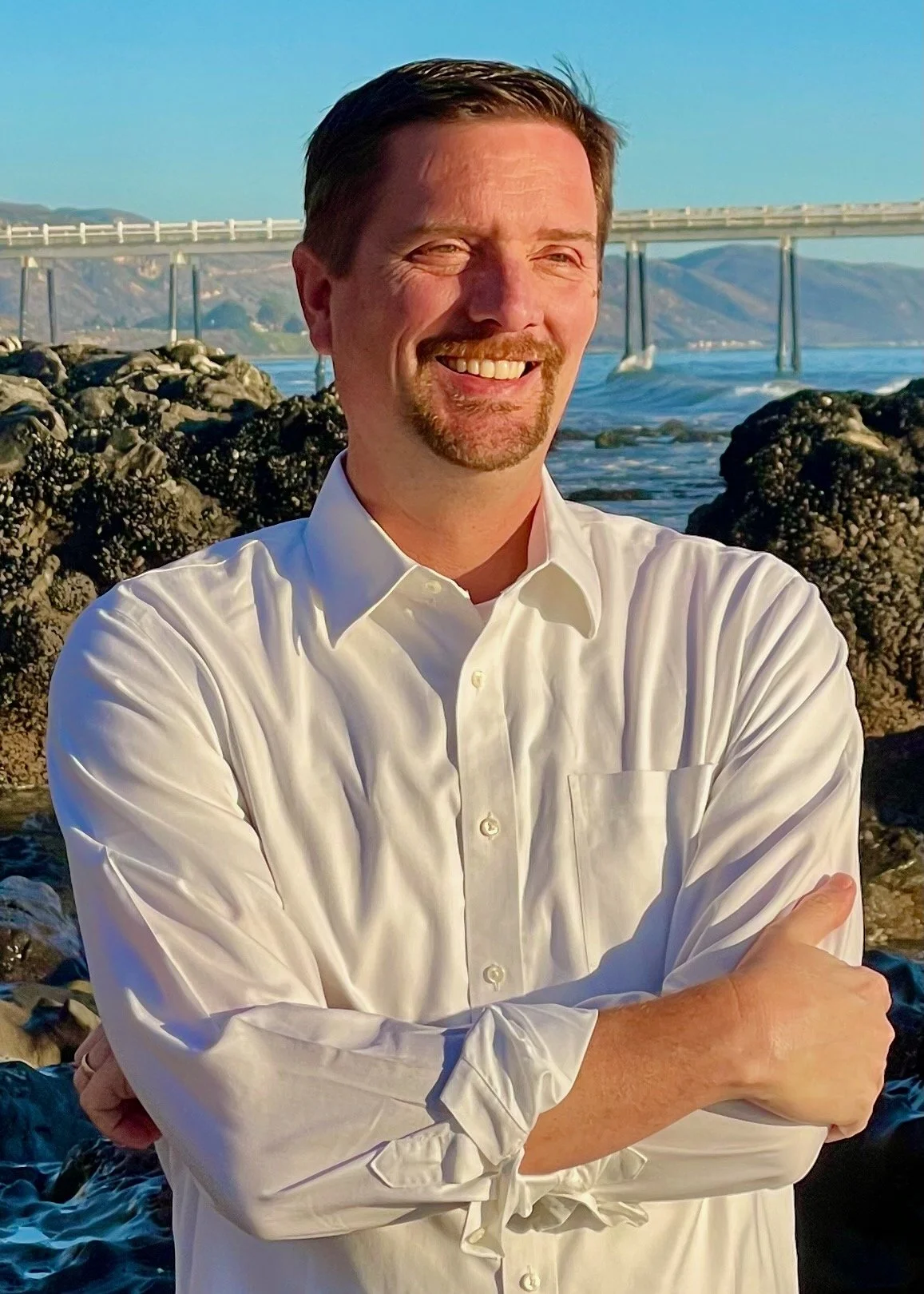 A man with dark hair and a beard, wearing a white button-up shirt, smiling with crossed arms, standing by the rocks near the ocean during sunset.