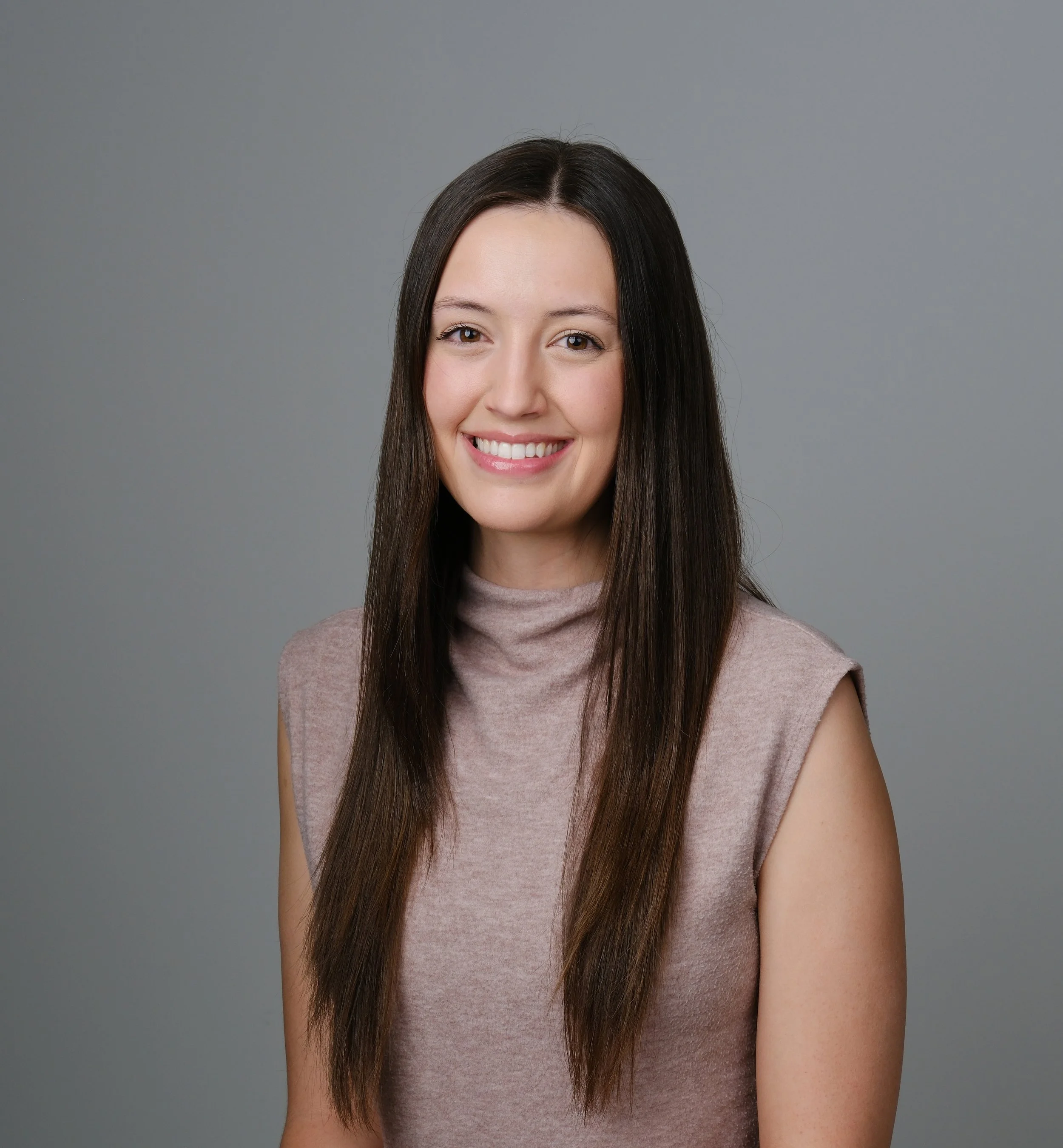 A smiling female therapist with long brown hair wearing a beige top against a grey background.