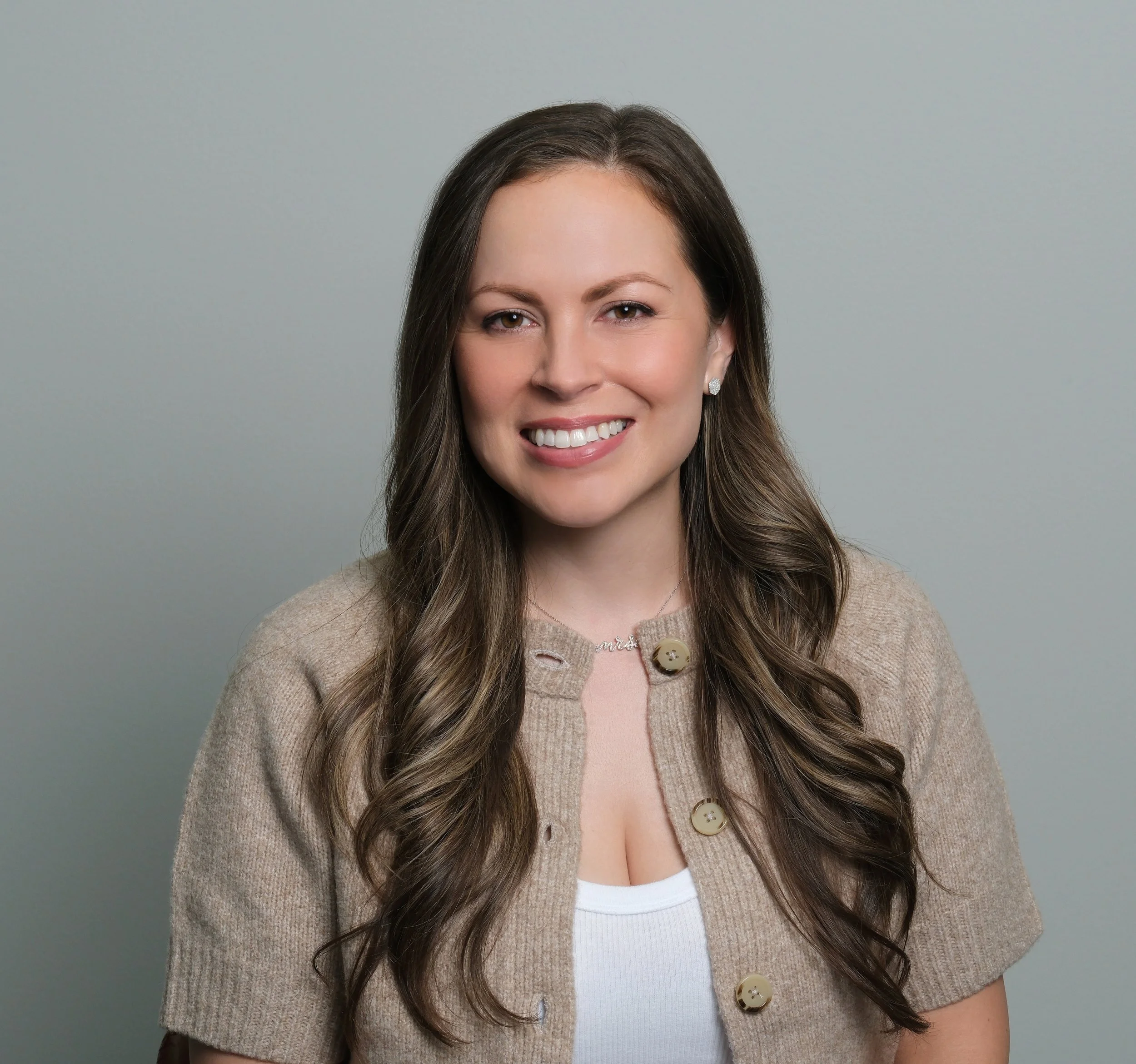 Smiling woman with long brown hair wearing a navy blue blouse and a silver necklace, posing against a gray background who is a clinician specializing in OCD treatment.
