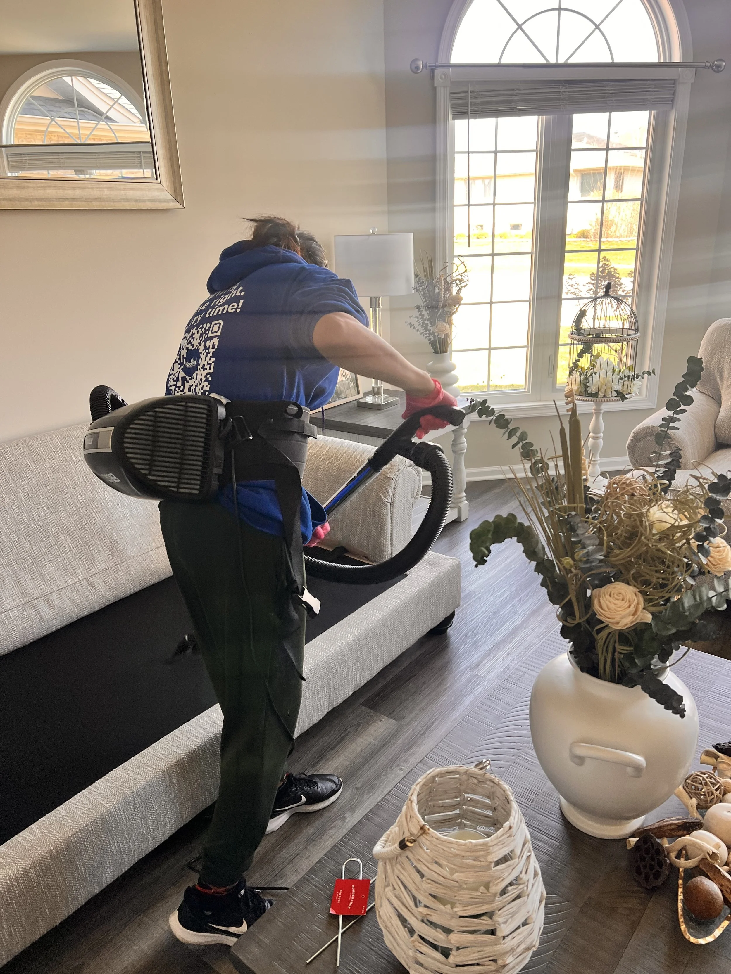 A person vacuuming a living room with sunlight streaming through large windows.