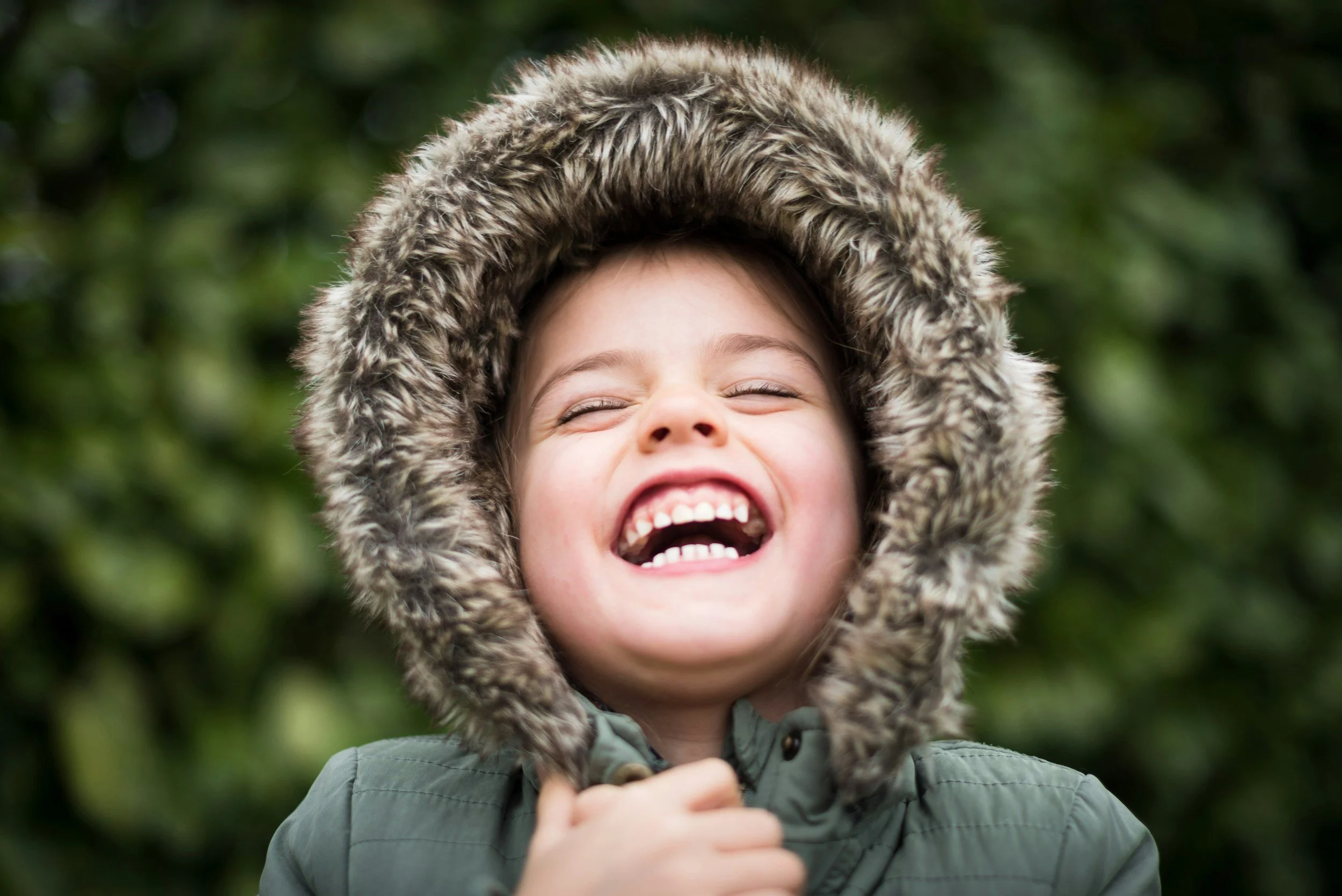 A young girl with a big smile, wearing a green jacket with a fur-lined hood, standing outdoors with green foliage in the background.