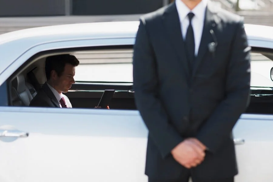 A man in a suit sitting in a car and looking at a tablet, with another man in a suit standing outside the car in front.
