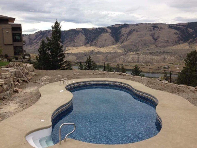 A scenic view of a curved swimming pool with a mountain landscape in the background. There are trees and a house to the left, with the pool surrounded by a light-colored concrete deck.