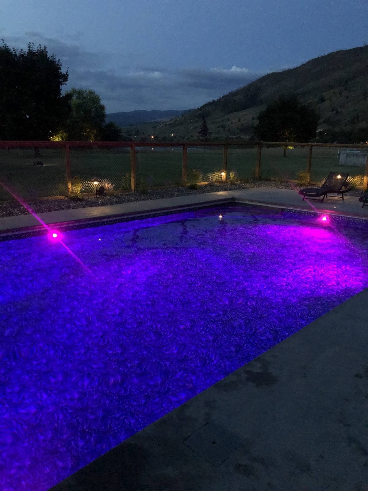 Swimming pool with purple lights at dusk, surrounded by a fence and chairs, with a scenic mountain and sky backdrop.