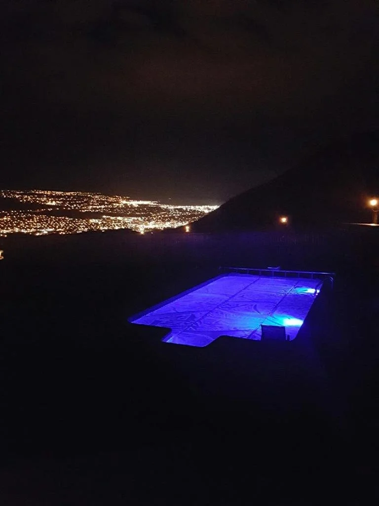 A pool illuminated with blue lights at night, overlooking a city skyline with twinkling city lights in the distance.