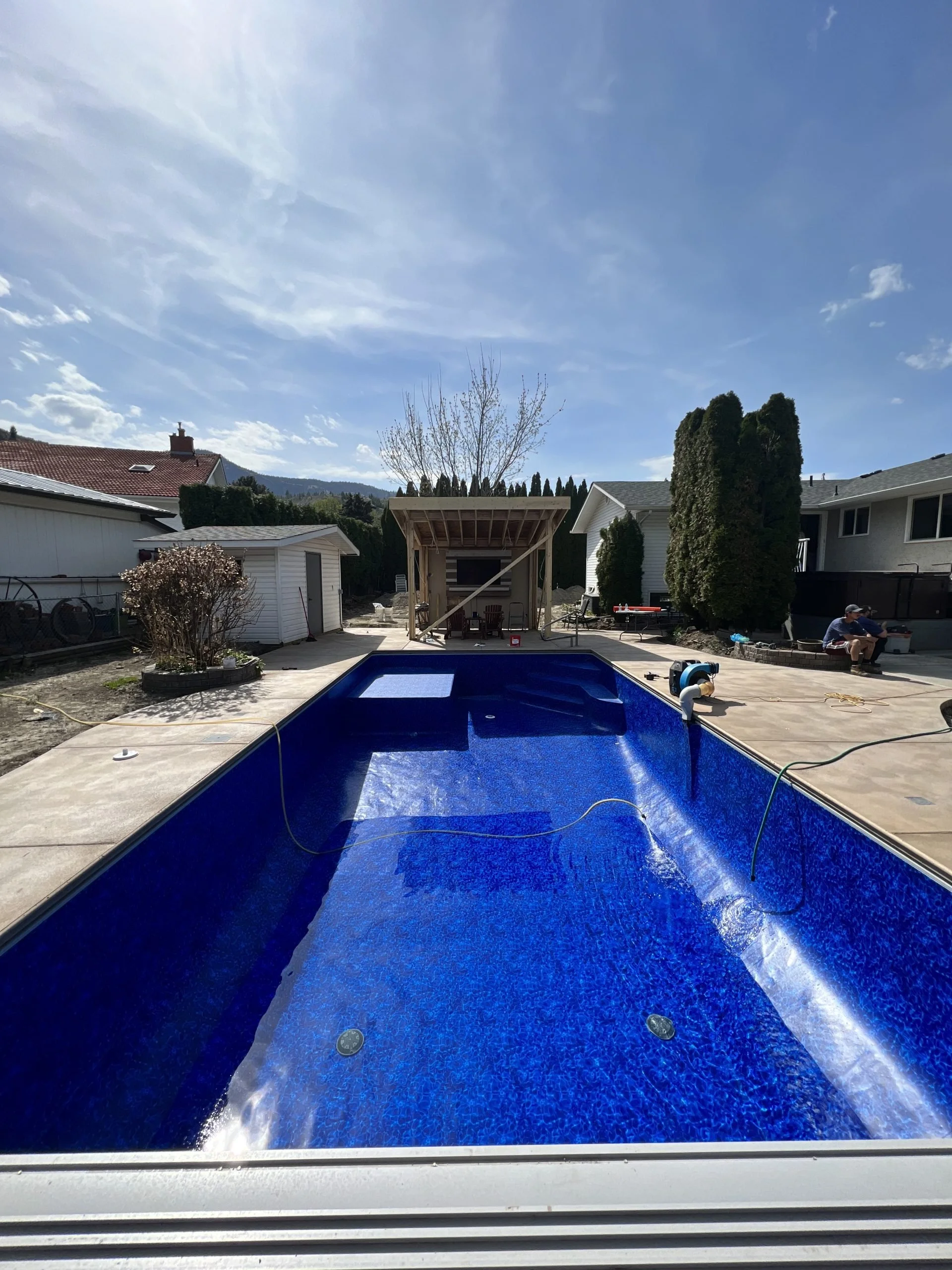 Empty swimming pool in backyard with patio area and shed, under a blue sky.