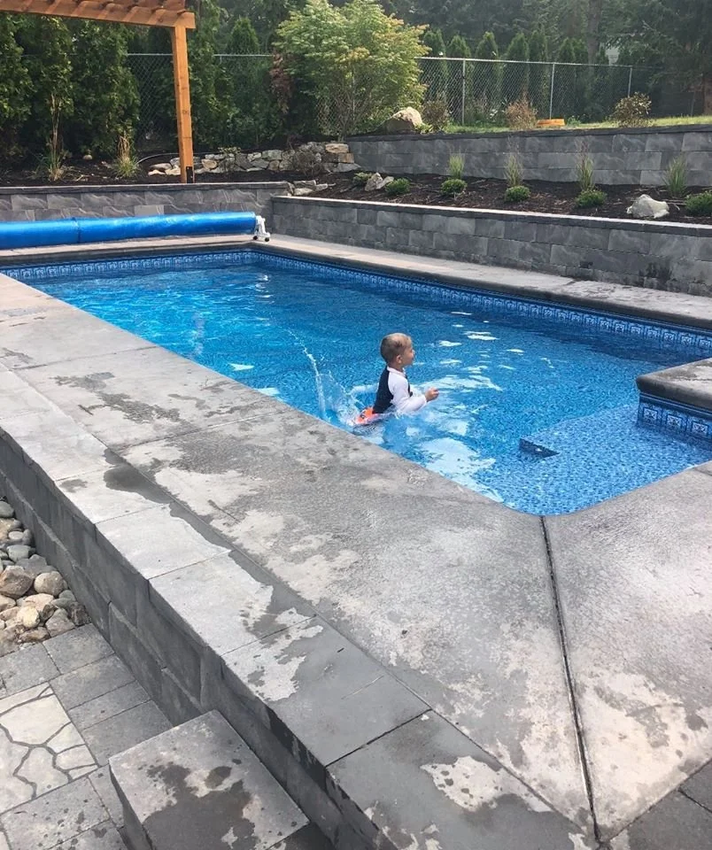 Child swimming in a backyard pool with gray stone patio and surrounding greenery.