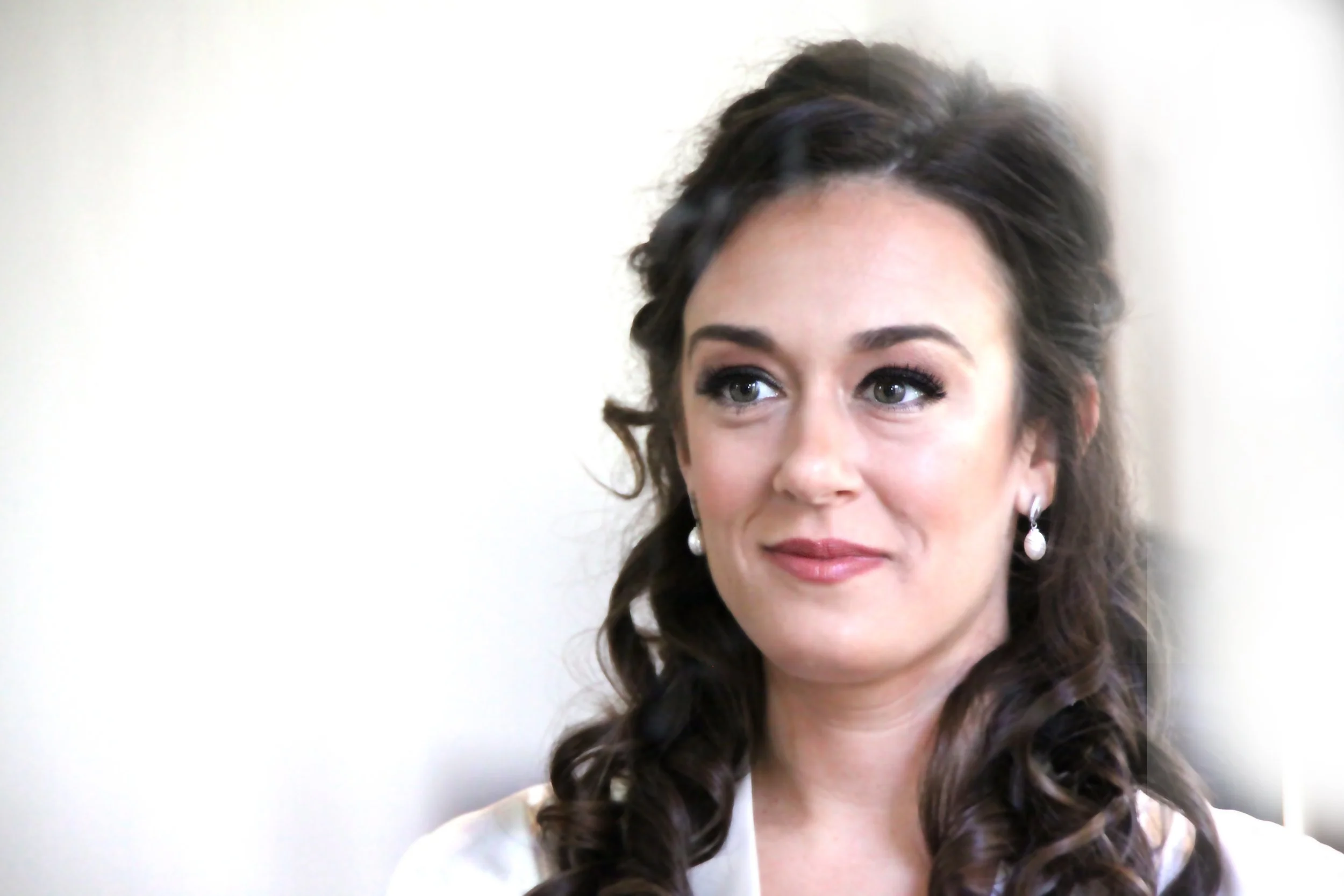 Close-up of a woman with dark curly hair, pearl earrings, and makeup, looking slightly to the right with a subtle smile.