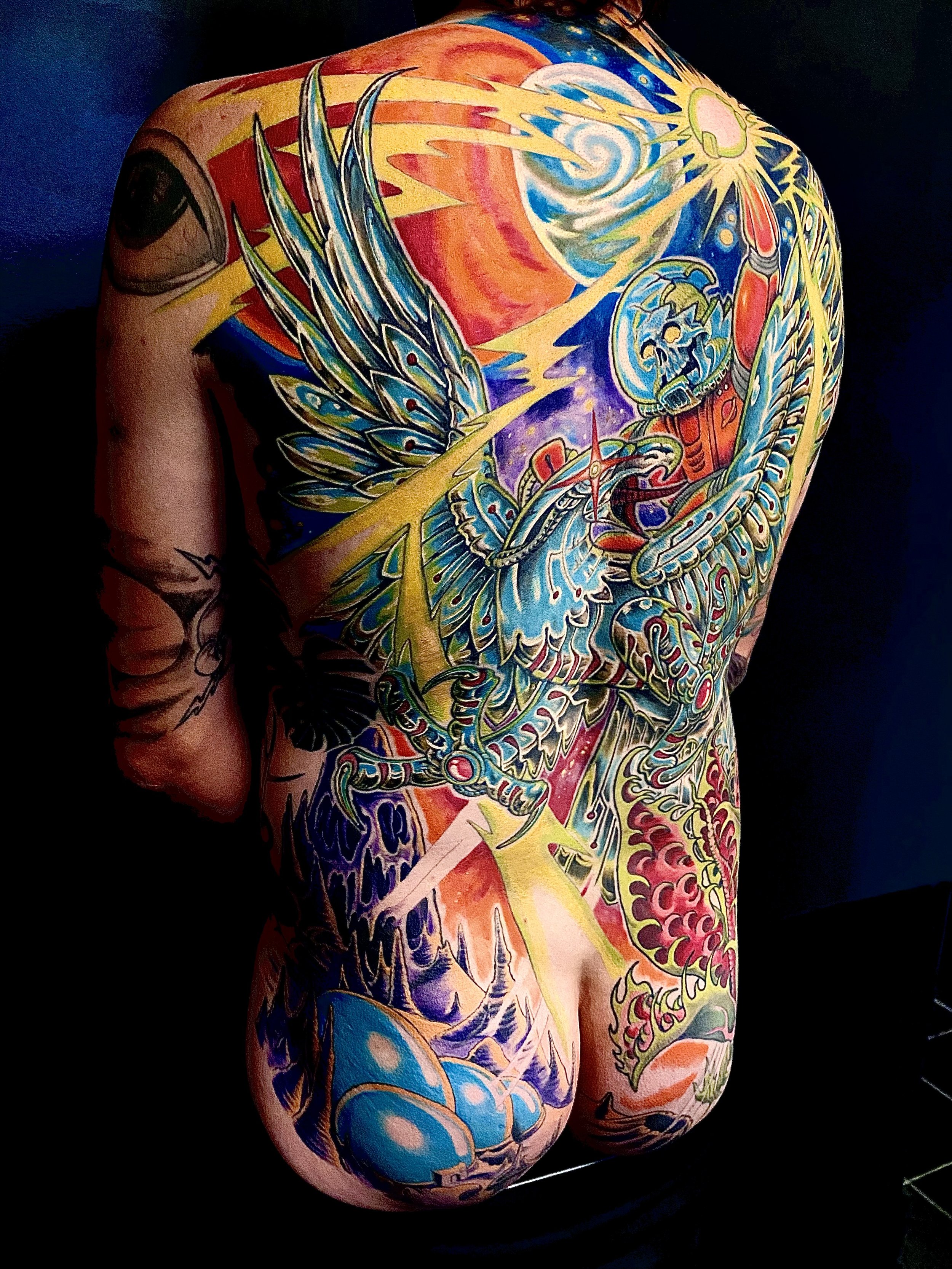 Colorful tattoo covering a person's back, featuring a dragon, phoenix, butterfly, skull, and celestial elements, with vibrant shades of blue, red, yellow, and purple.