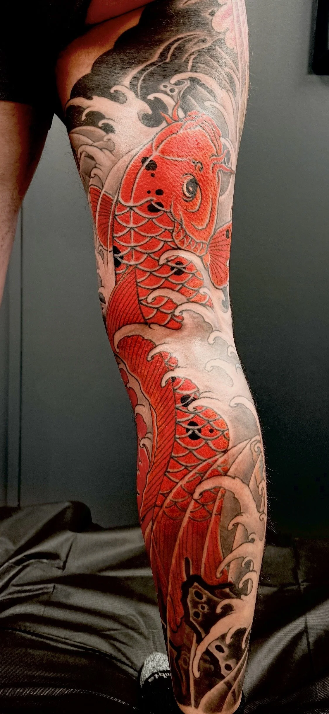 Tattoo of a red koi fish swimming amongst stylized waves and clouds on a person's leg.