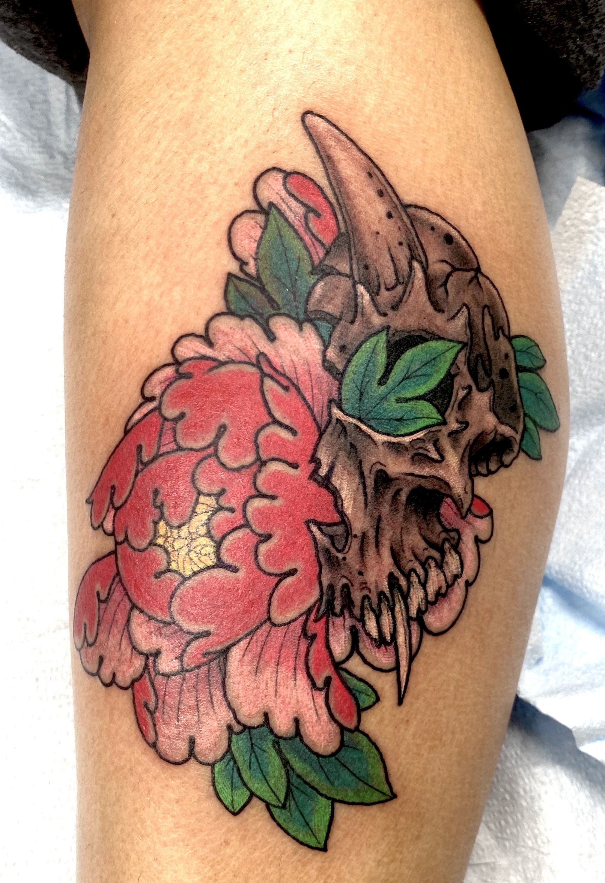 Tattoo of a skeleton skull with sharp teeth and hollow eye sockets, surrounded by a large pink flower with green leaves.