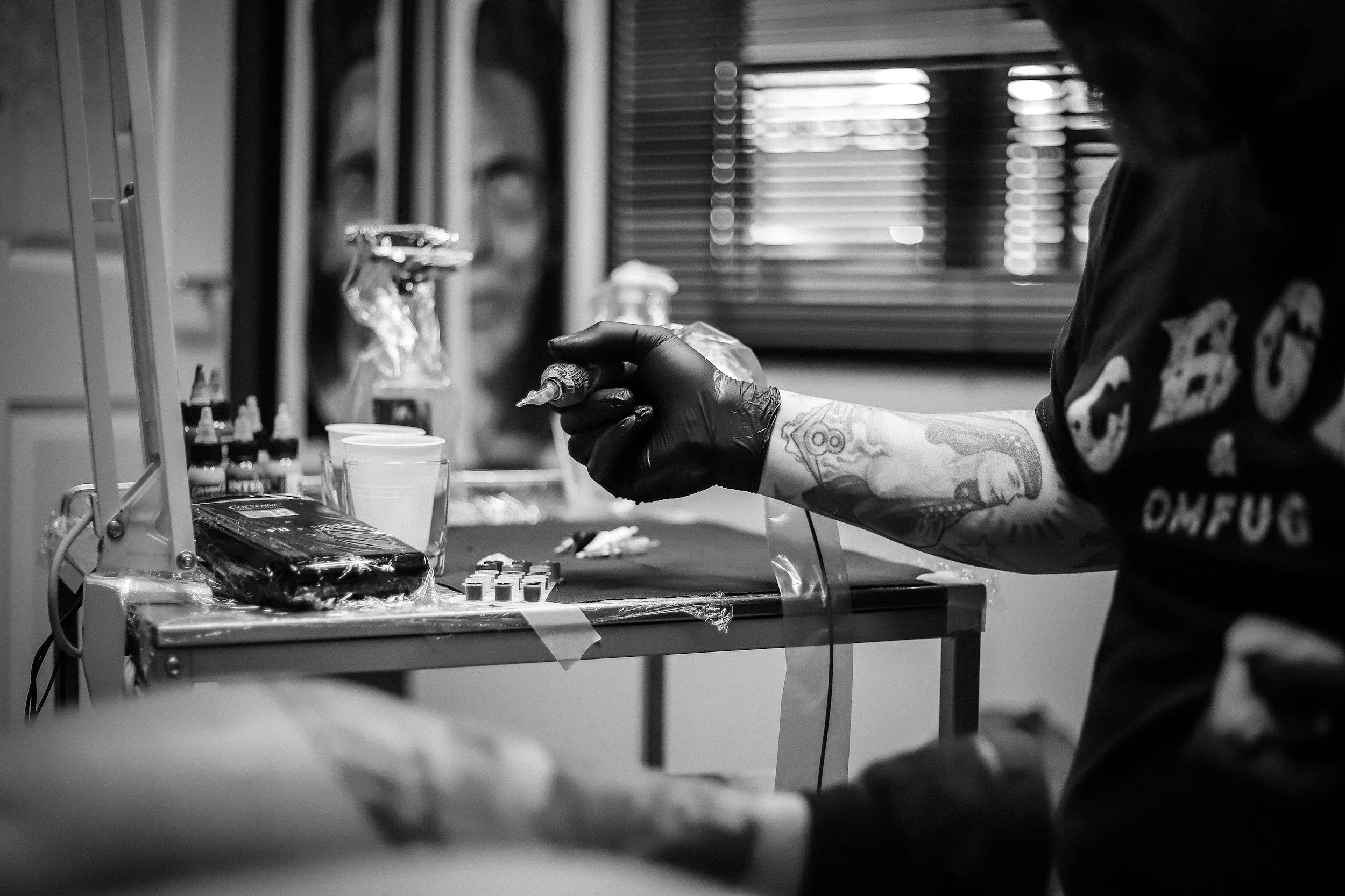 Tattooed person wearing gloves preparing tattoo ink in a tattoo studio, with tattoo supplies on a table and an artistic portrait in the background.