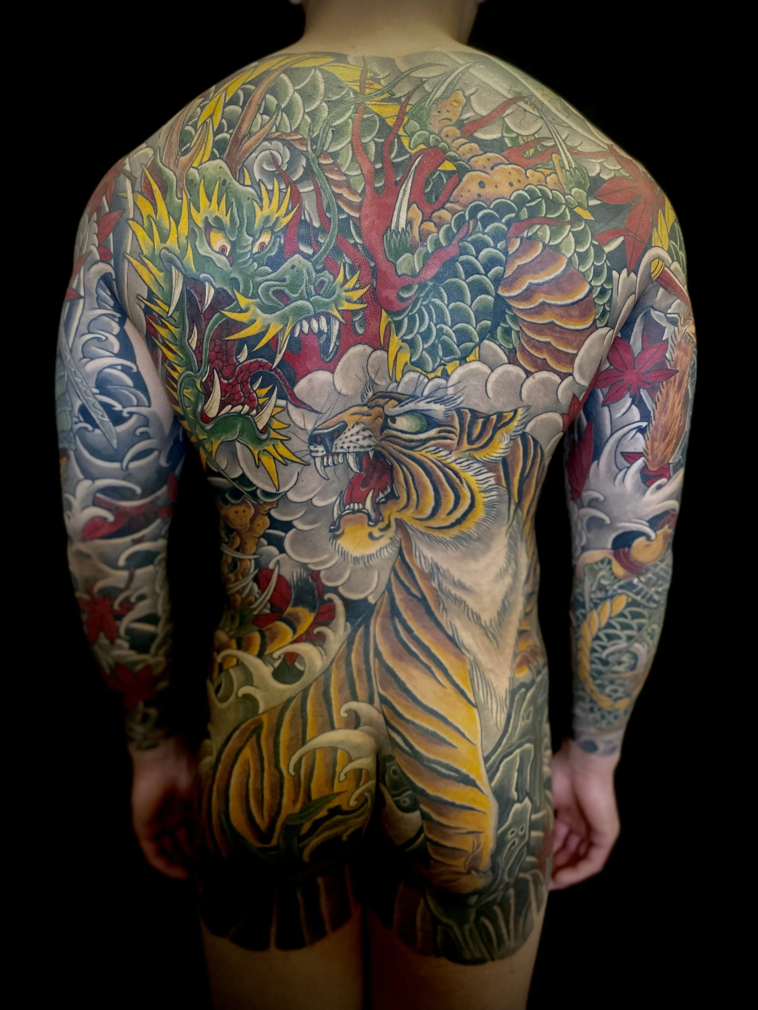 Full back and arms tattoo of traditional Japanese style featuring a fierce green dragon, a roaring tiger, a red maple leaf, and swirling clouds.