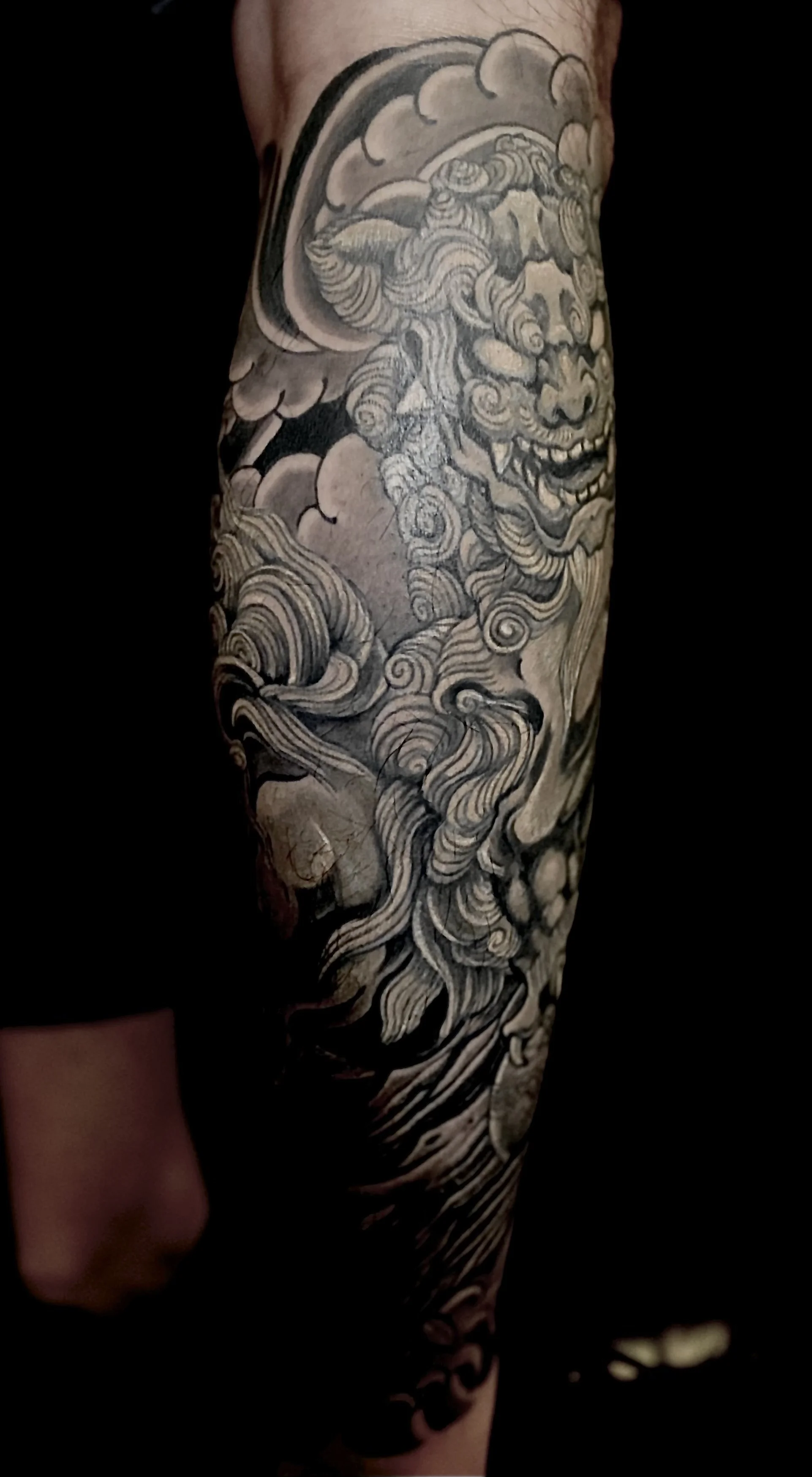 Black and gray Japanese-style tattoo sleeve featuring a fierce mythical lion or dragon face, swirling clouds, and waves.