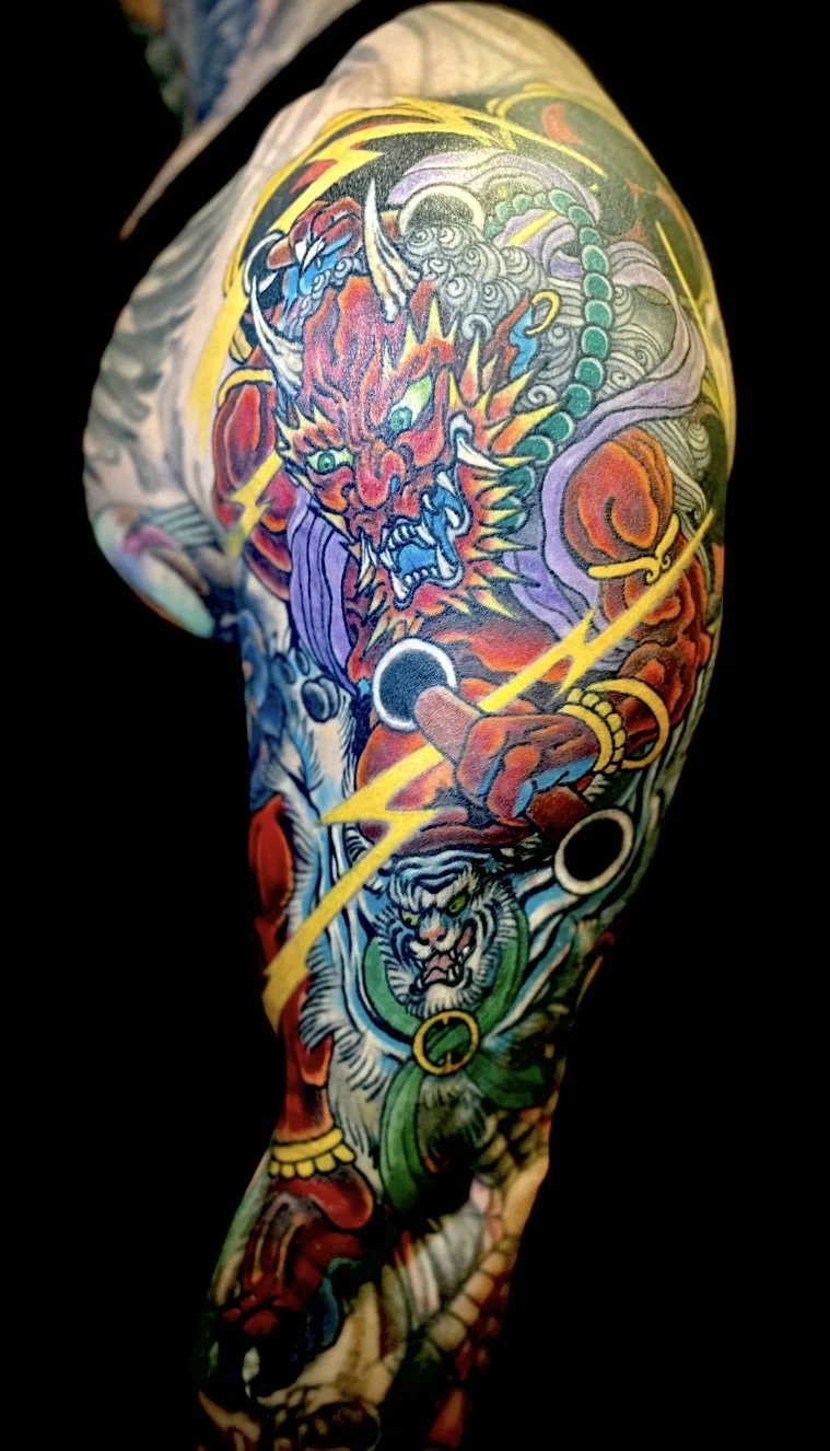 Colorful tattoo sleeve featuring a fierce mythical creature with a red face, horns, and sharp teeth, wielding lightning bolts, and a tiger with green eyes and sharp teeth at the wrist.