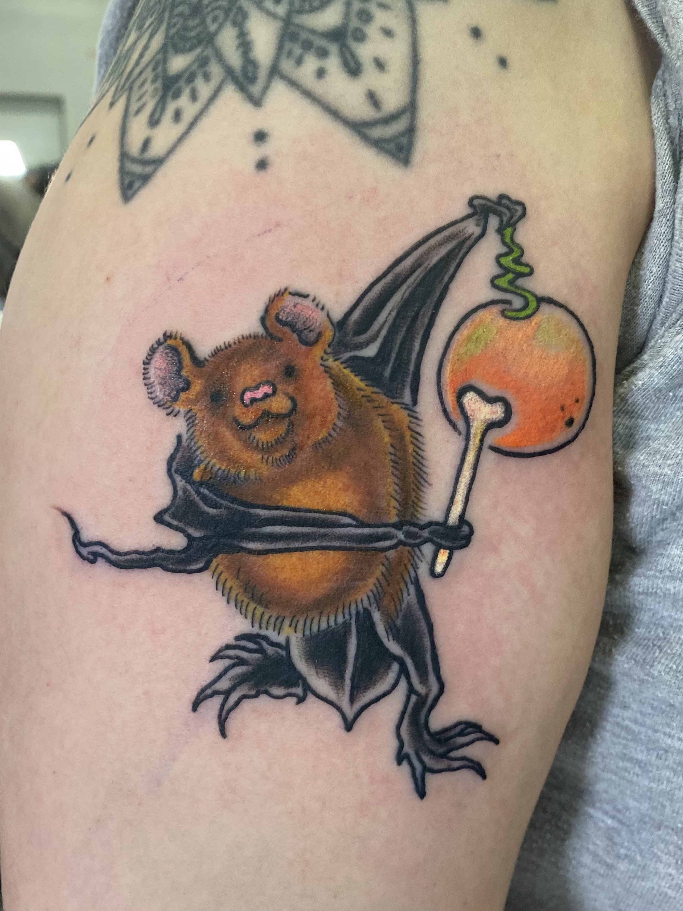 A tattoo of a bat with bear-like face holding a balloon and a stick, with a colorful design and cartoonish style.
