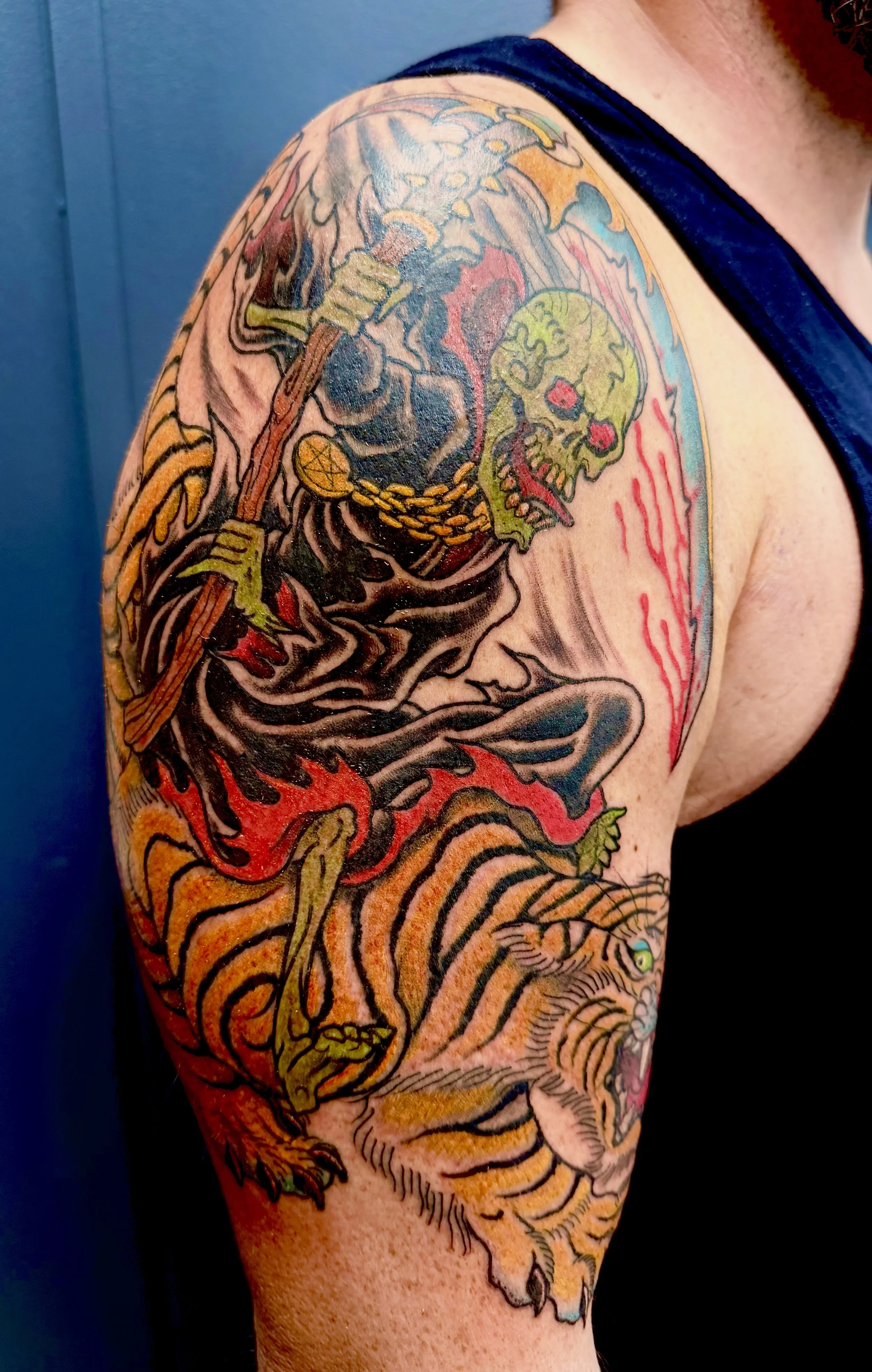 Tattoo of a fierce tiger with a skull in its mouth and a chain in its mouth on person’s upper arm and shoulder.