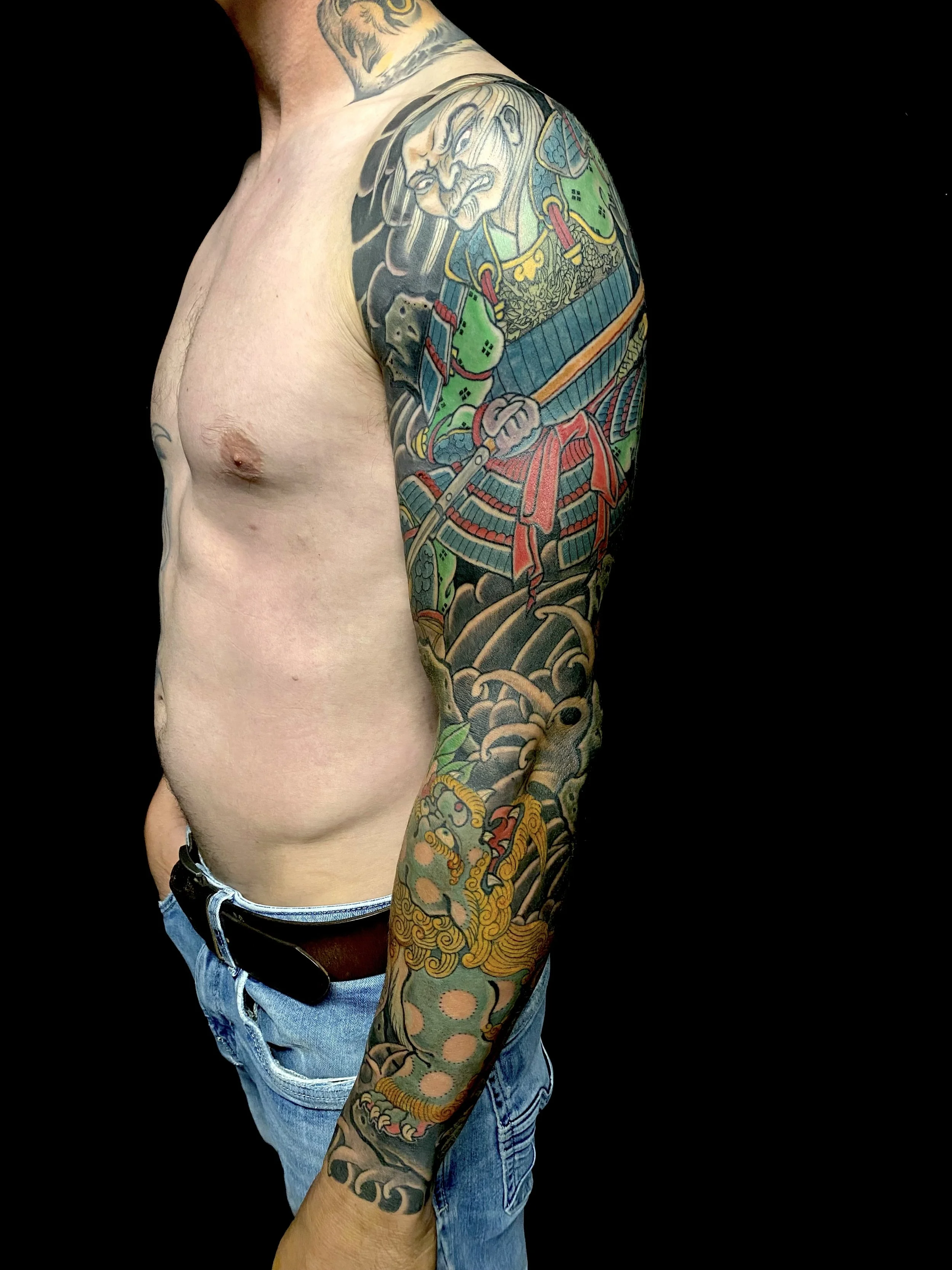 A man with extensive colorful Japanese-style tattoos covering his upper arm and shoulder, featuring a fierce warrior, a dragon, and traditional motifs, against a black background.